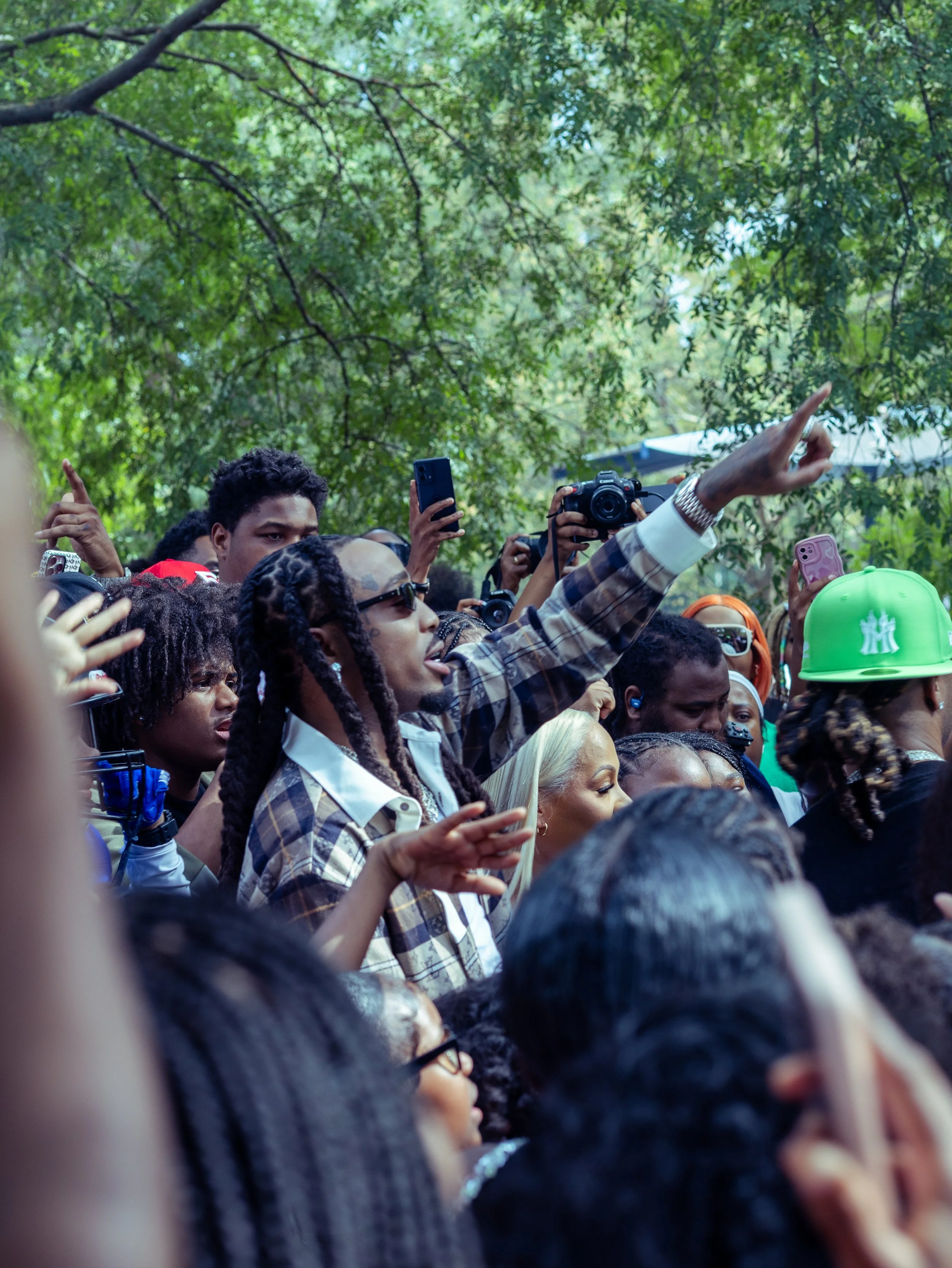 Crowd of people at an outdoor event, some taking photos or recording with smartphones and cameras, with a person in a plaid shirt and sunglasses raising one arm and pointing upward, surrounded by trees.