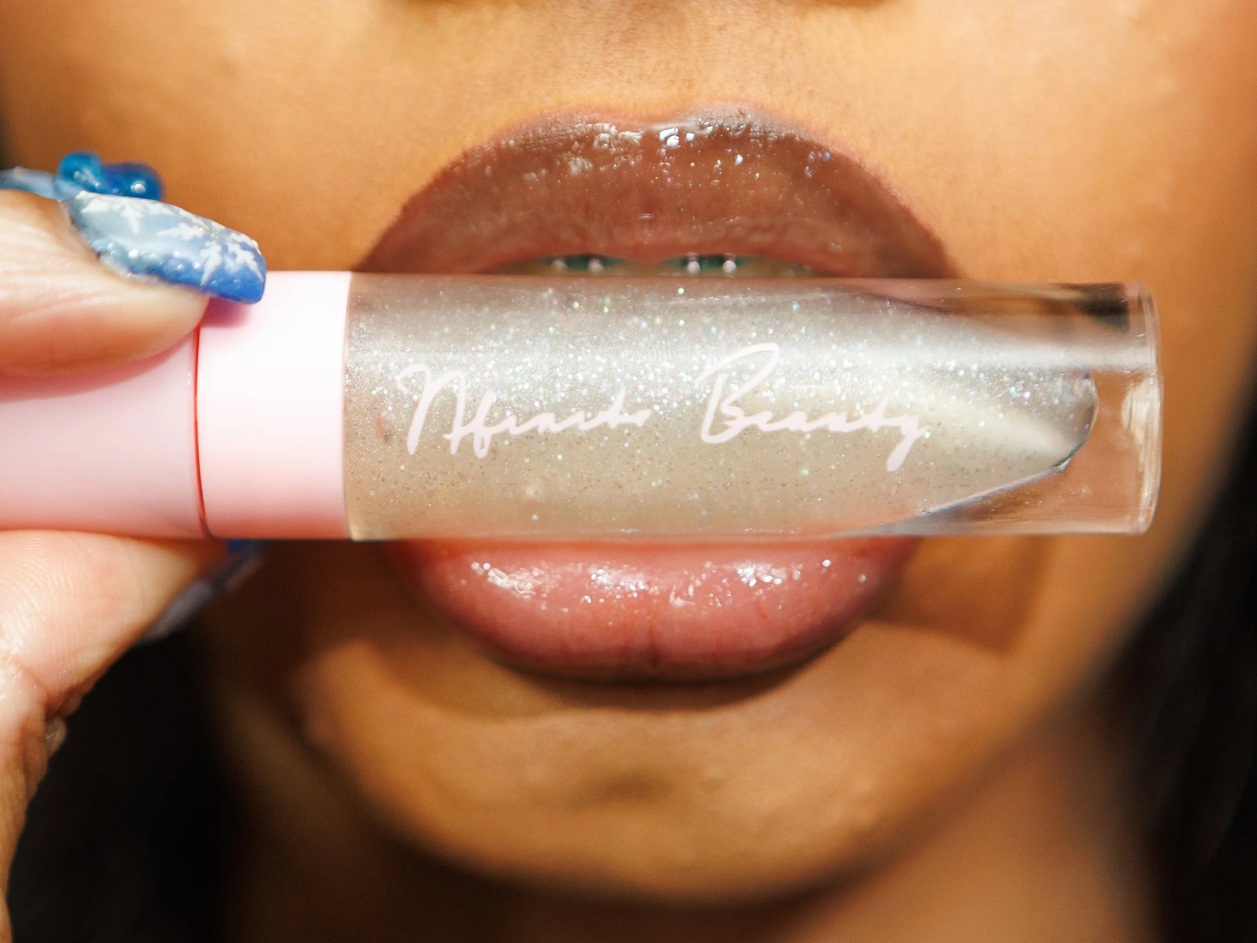 Close-up of a person with lips slightly parted holding a lip gloss tube labeled 'Thousand Beauty' against their lips, with blue nail polish visible.