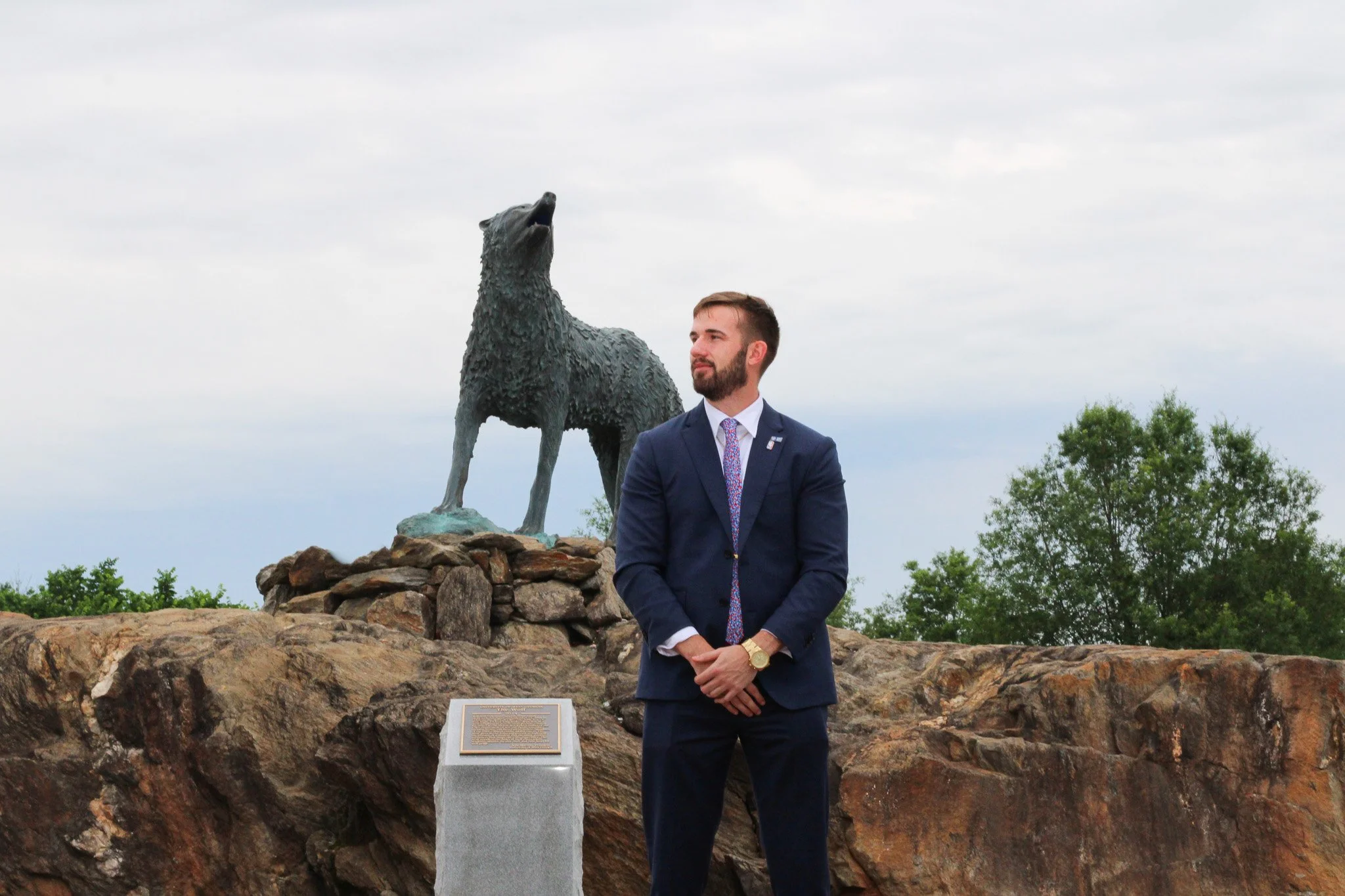 A man in a suit stands with hands clasped in front, in front of a monument of a howling wolf statue on a rocky base, with trees and cloudy sky in the background.