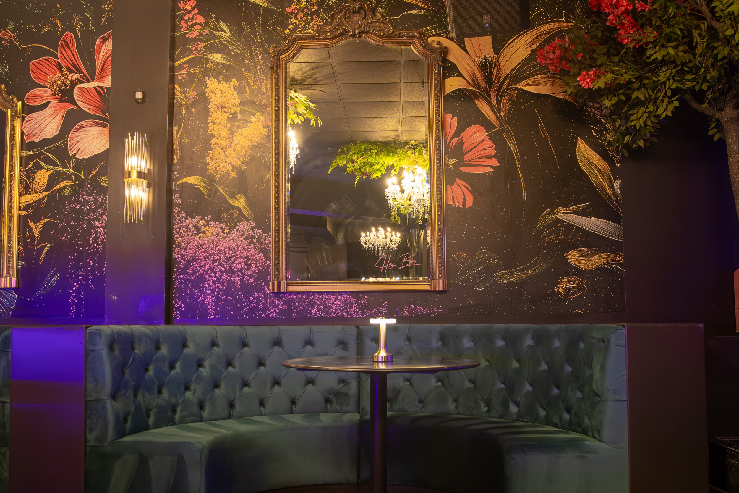 Elegant lounge with dark floral wallpaper, a large ornate mirror, plush velvet banquette seating, a small table with a lit candle, and chandeliers reflected in the mirror.