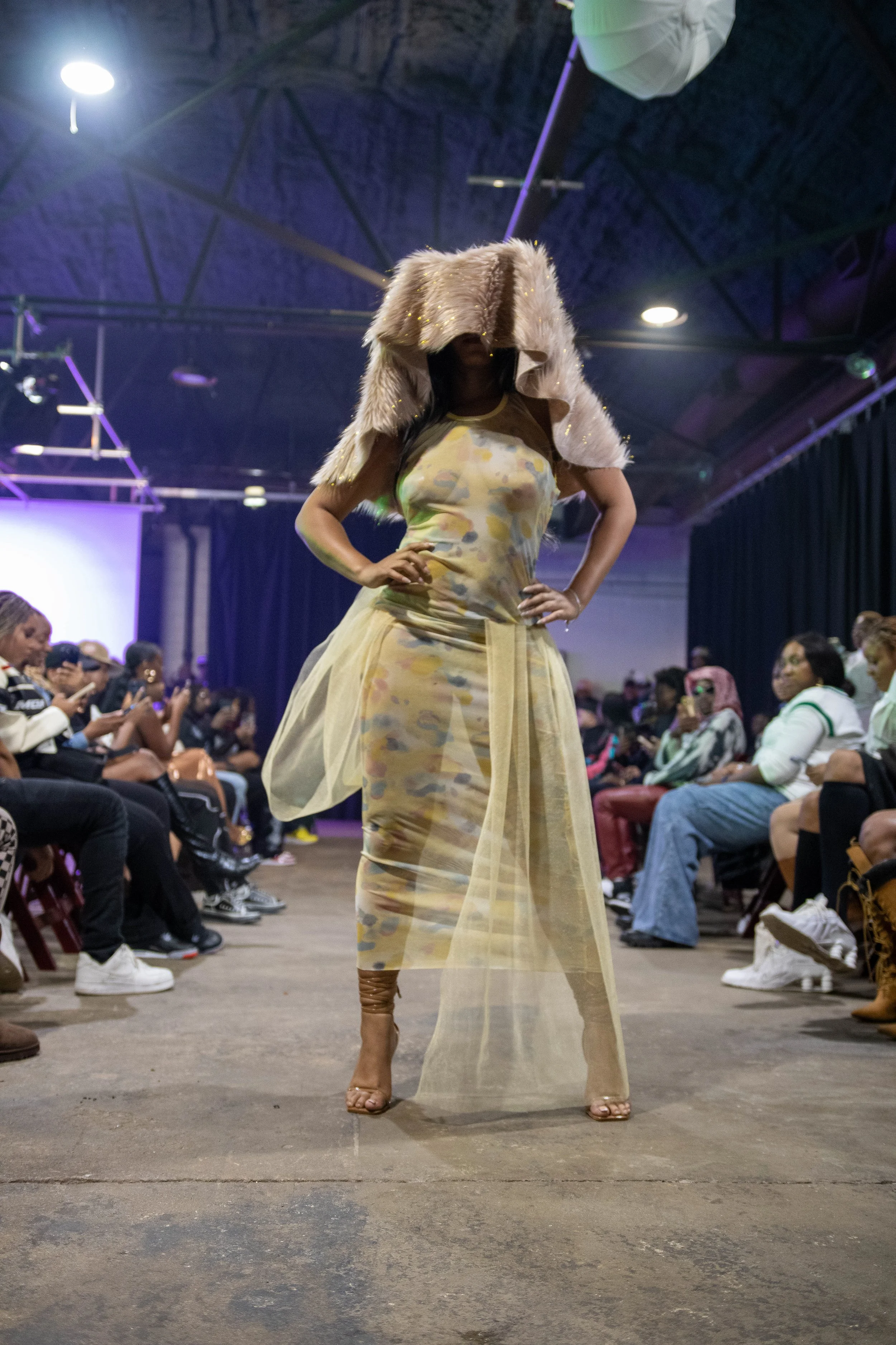 A fashion model walking down the runway wearing a multi-colored, see-through dress with a sheer yellow overlay and a fur coat-like accessory on her shoulders. Audience members are seated on either side of the runway in a large indoor venue.