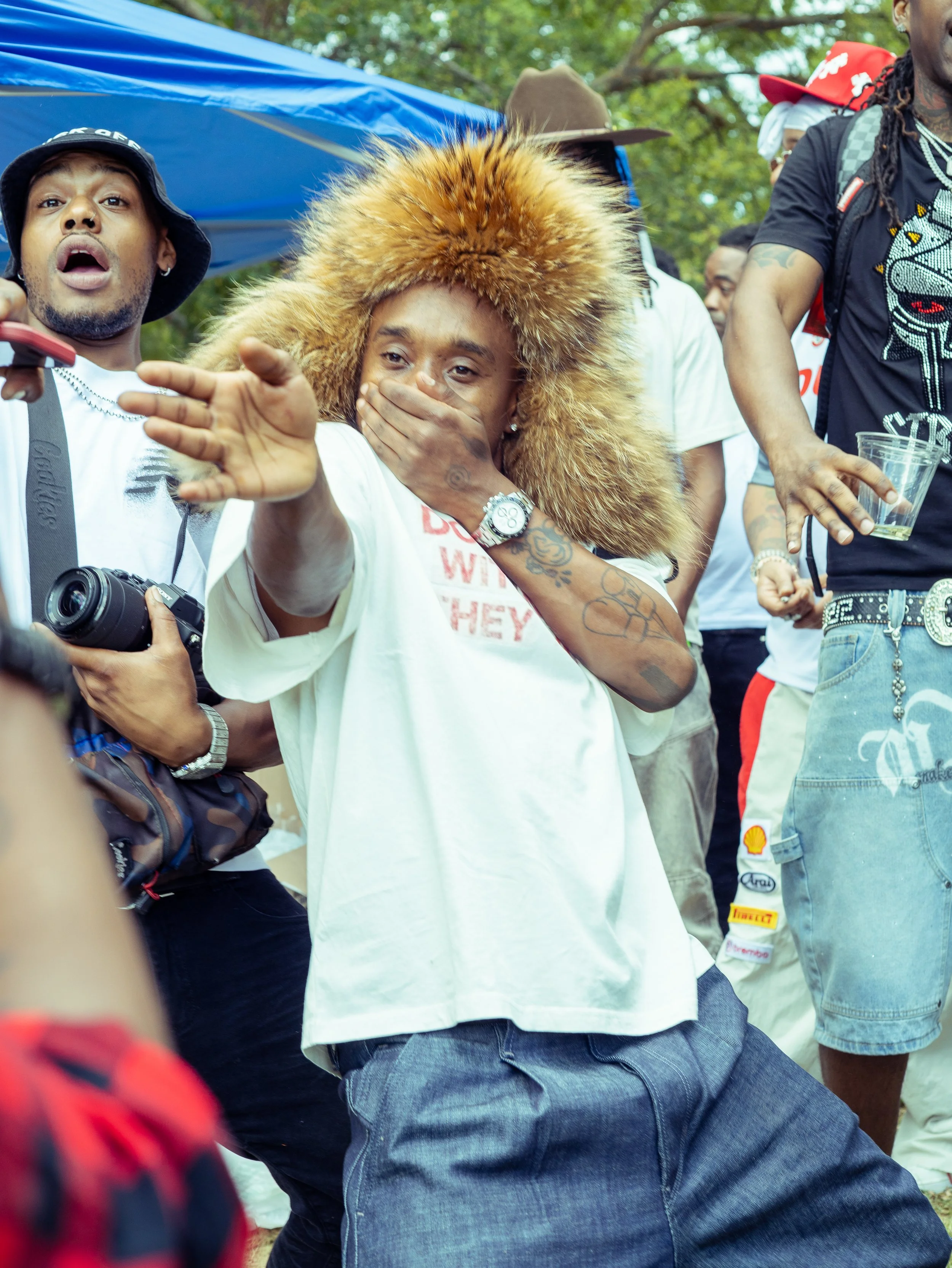 Group of people outdoors, with one woman wearing a large fur hat covering her head, holding her hand to her mouth, and appearing surprised or emotional.