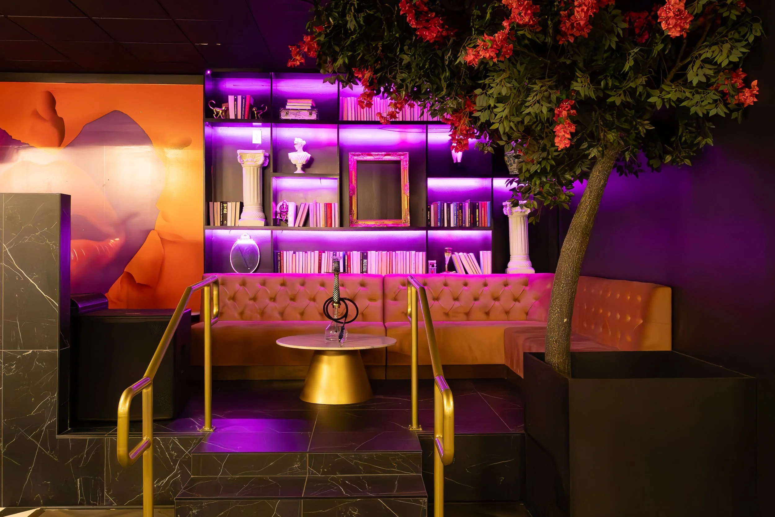 A cozy indoor lounge with a pink tufted sofa, a small round white table, and a hookah in the center. Behind the sofa, there's a bookshelf with purple lighting, decorated with books, white classical busts, and a framed mirror. To the right, a large po