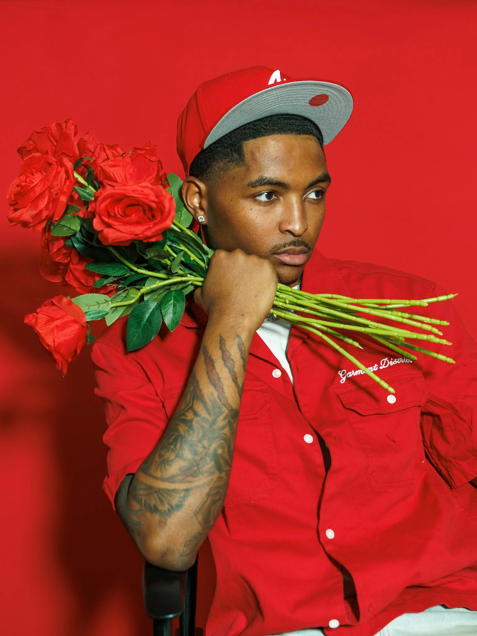 A young man with tattoos on his arm holding a bouquet of red roses on his shoulder, wearing a red shirt and a red and gray cap, against a red background.