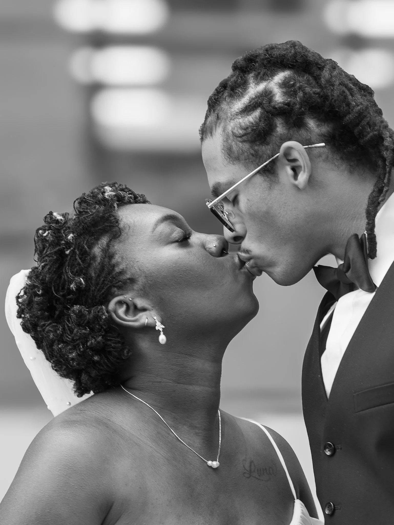 A couple in formal attire sharing a kiss, with the woman wearing jewelry and the man wearing a tuxedo and glasses, in a black-and-white photograph.