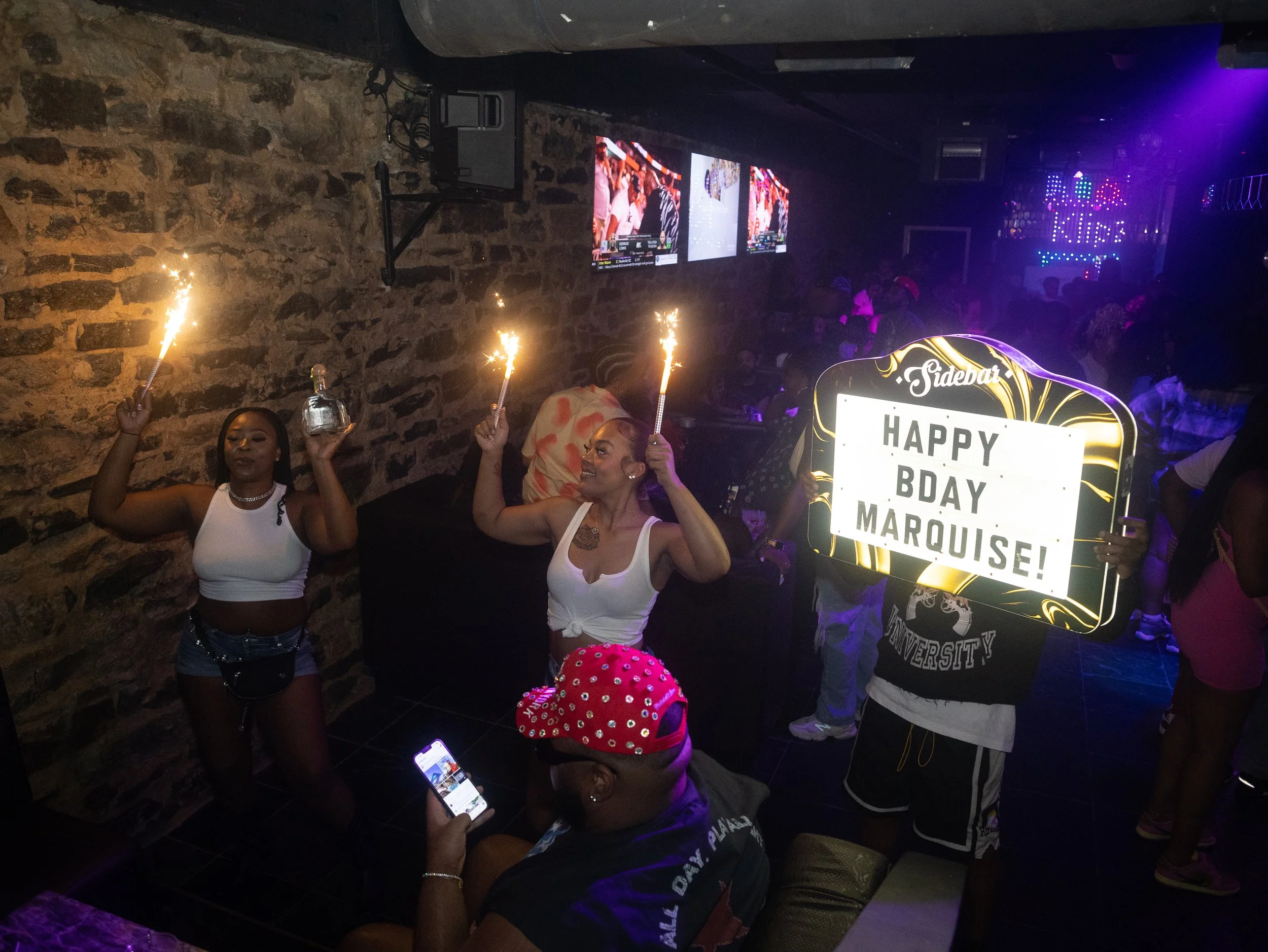 Group of women celebrating a birthday with sparklers in a dimly lit bar or club, one holding a bottle and another holding a sign that reads 'HAPPY BDAY MARQUISE!'