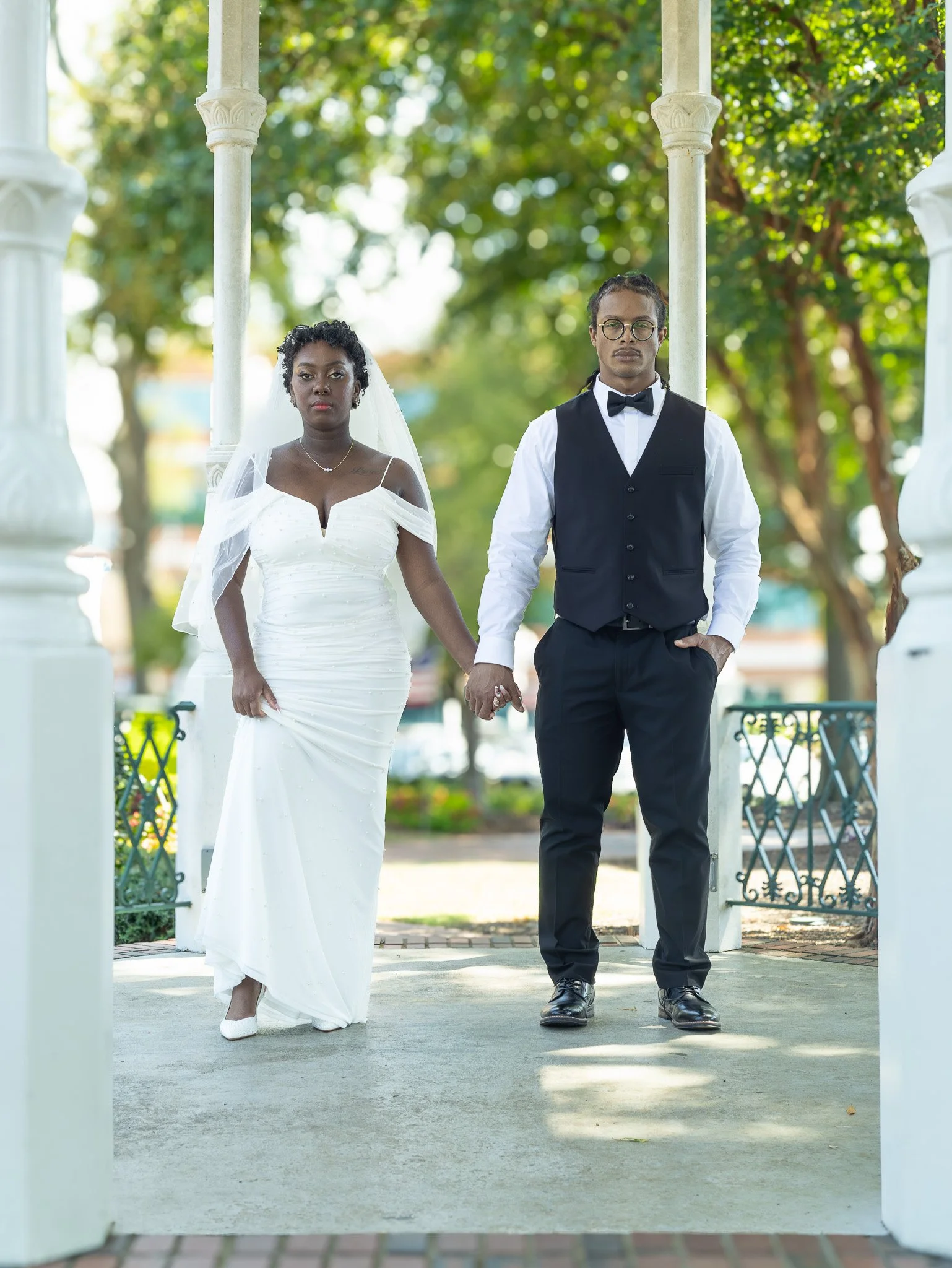 A bride and groom standing hand in hand outdoors, under a white gazebo with green trees in the background. The bride is wearing a white wedding dress with off-the-shoulder sleeves, and the groom is dressed in a black vest, white shirt, black bow tie,