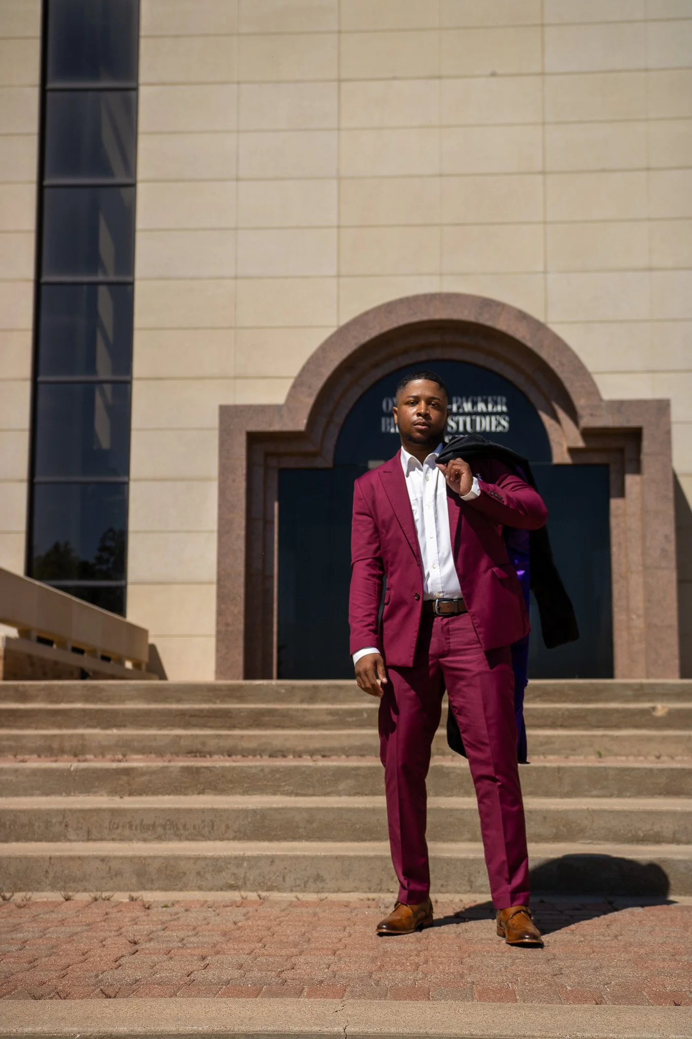 Young man in a burgundy suit standing on steps outside a university building holding a black coat over his shoulder.