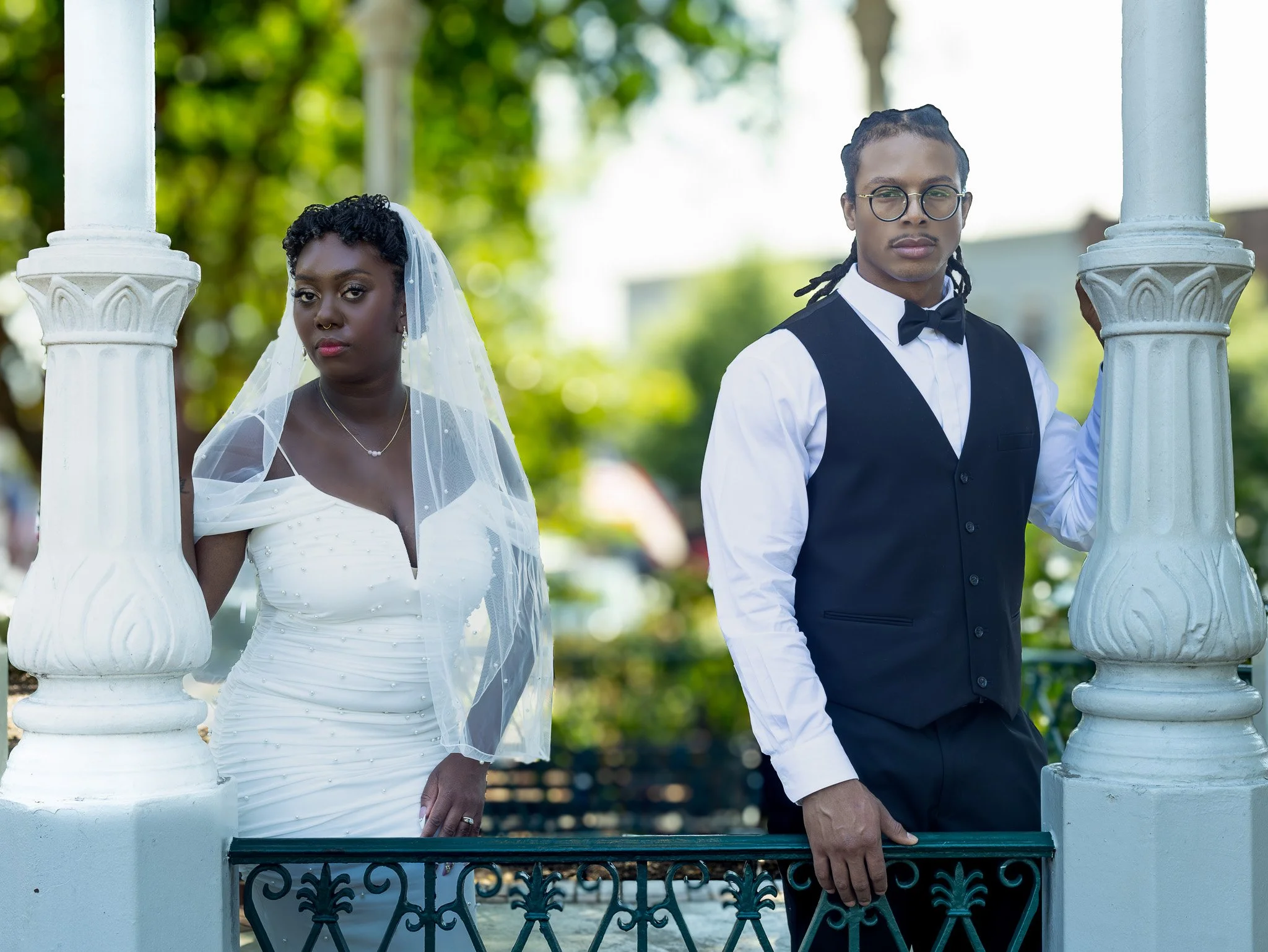 A bride in a white wedding dress and veil stands beside a groom in a black vest and bow tie, both leaning against white decorative pillars outdoors.