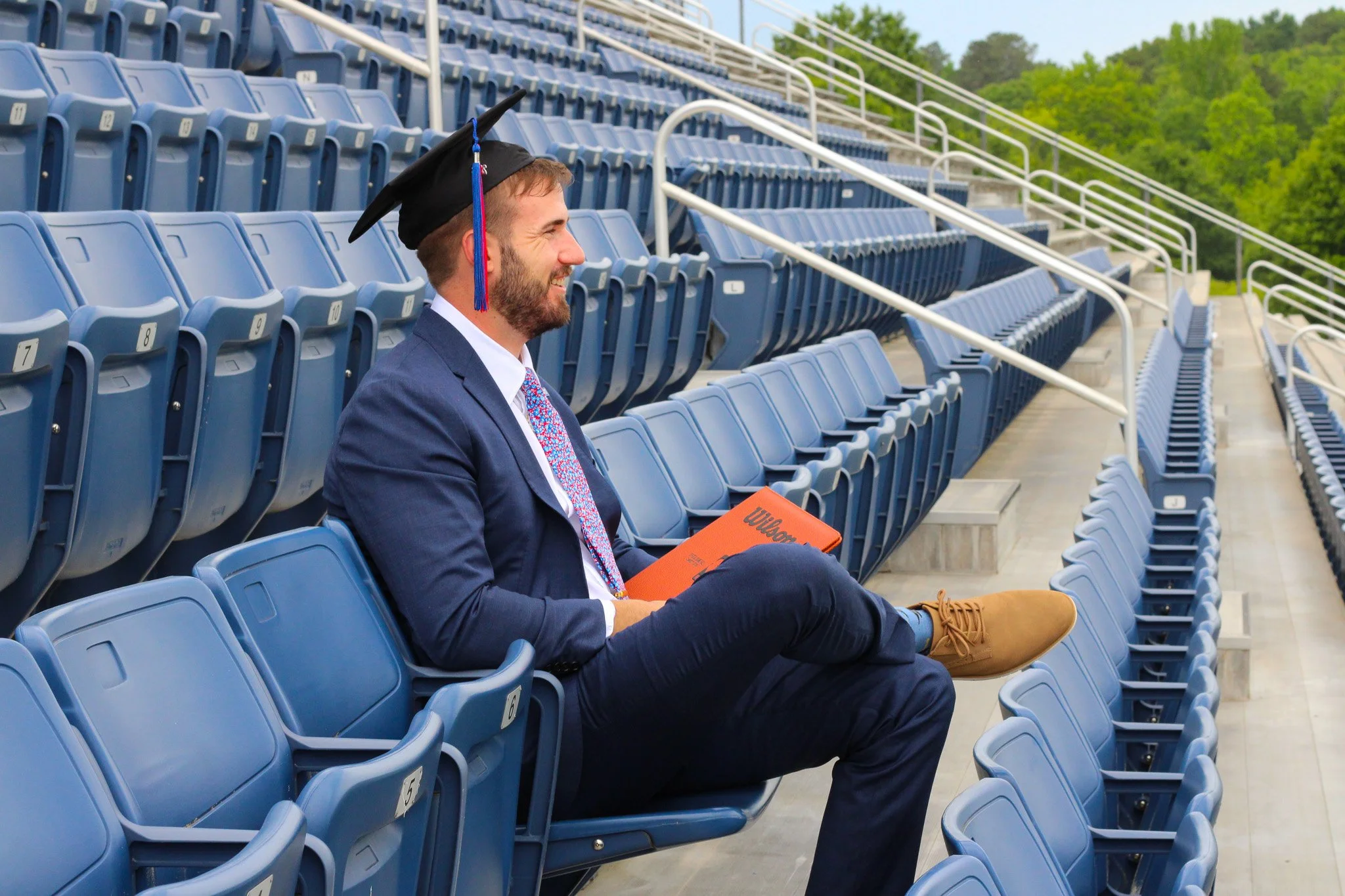 A man in a blue suit and graduation cap sitting alone in an empty outdoor stadium, smiling and holding a clipboard.