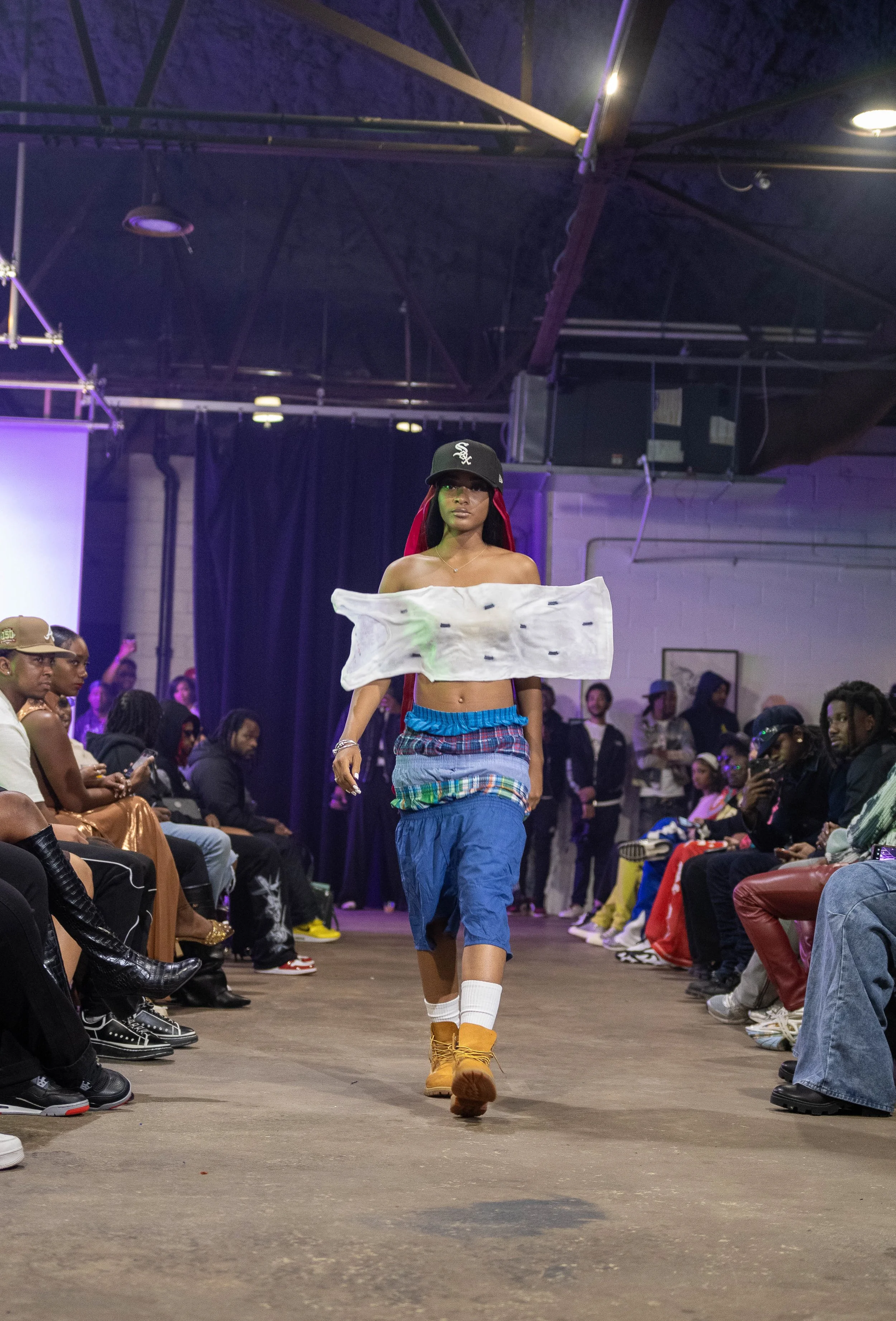 A fashion runway model is walking down the runway with an audience on both sides. The model wears a black cap, off-shoulder top cover with a patchwork skirt, yellow boots, and white socks.