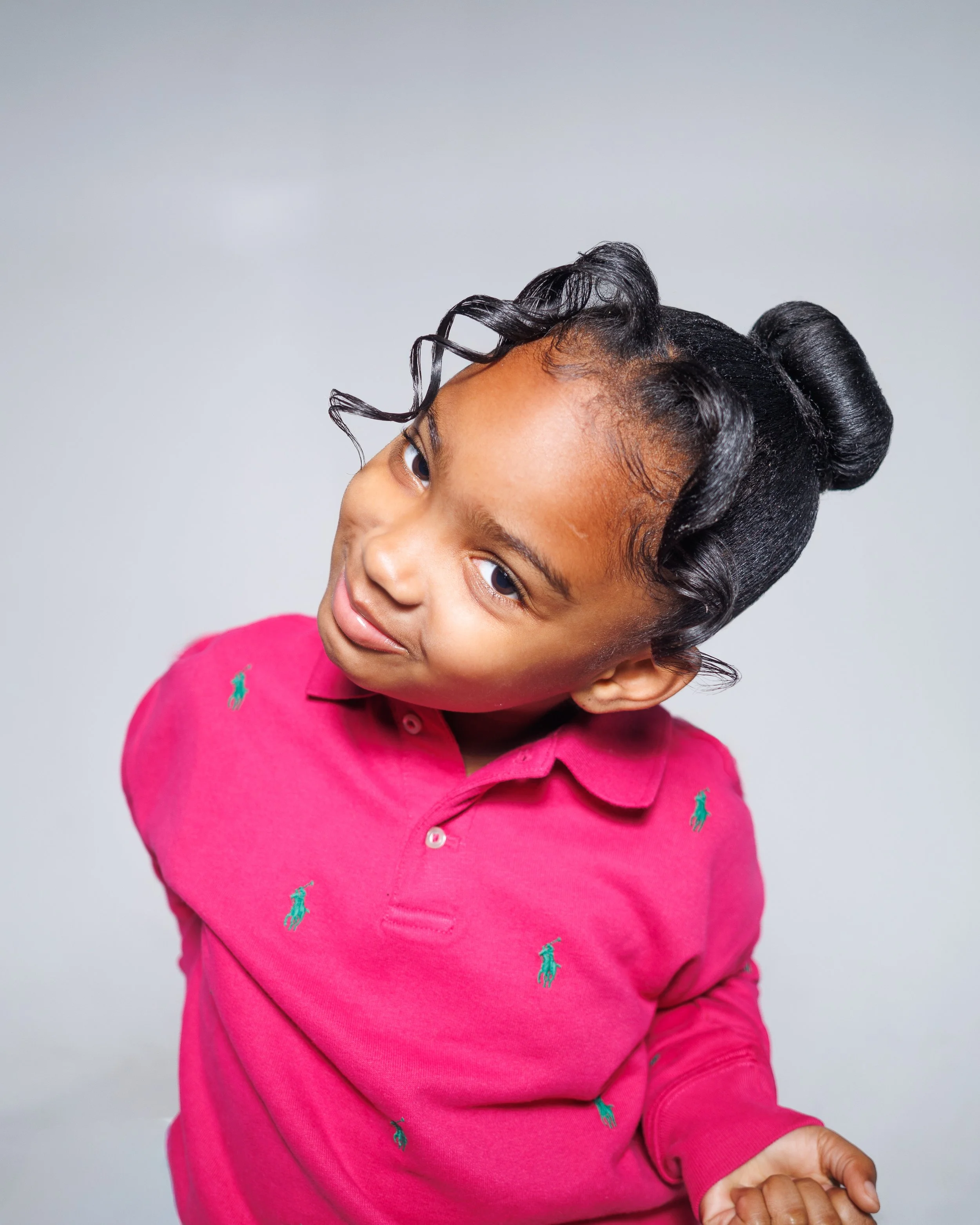 A young girl with styled curly hair in buns and loose curls, wearing a bright pink polo shirt with green embroidered Polo Ralph Lauren logos, looking at the camera with a playful expression against a plain light gray background.