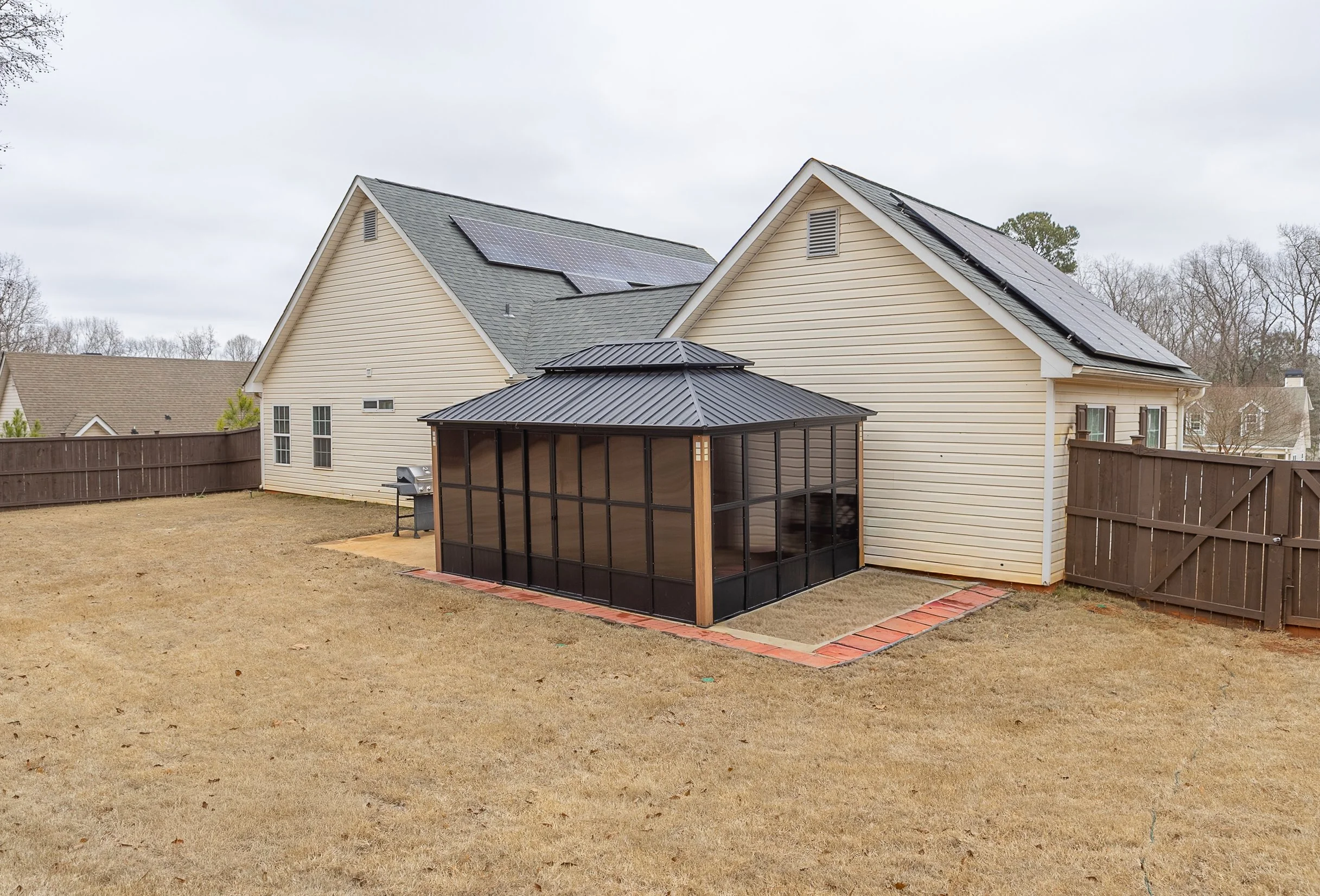 Backyard with two beige houses, one with solar panels on the roof, and a screened porch with a black metal roof, brick border, and concrete slab, surrounded by a wooden fence, on a cloudy day.