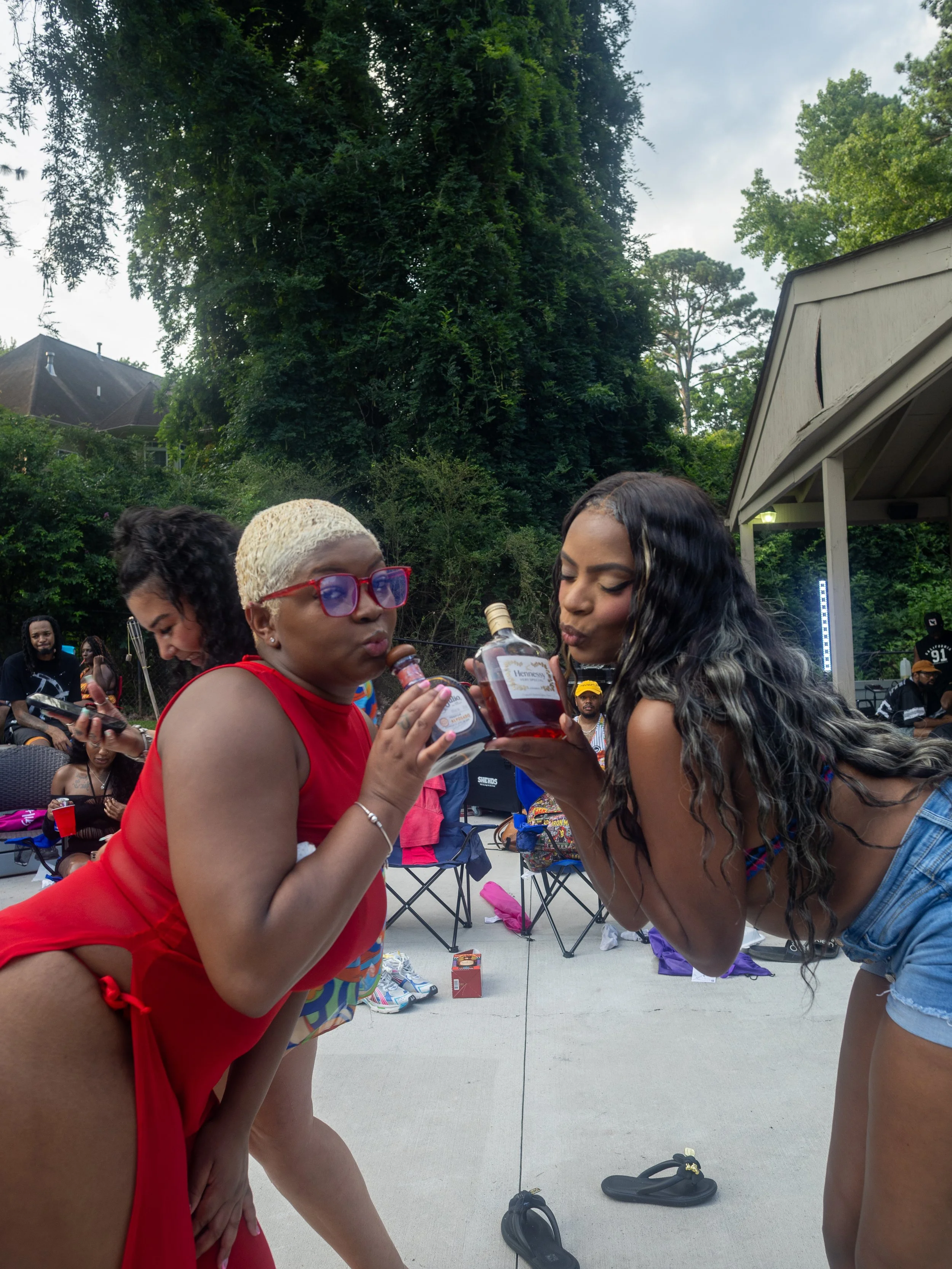 Two women are holding liquor bottles and pretending to drink from them at an outdoor gathering, with people and trees in the background.