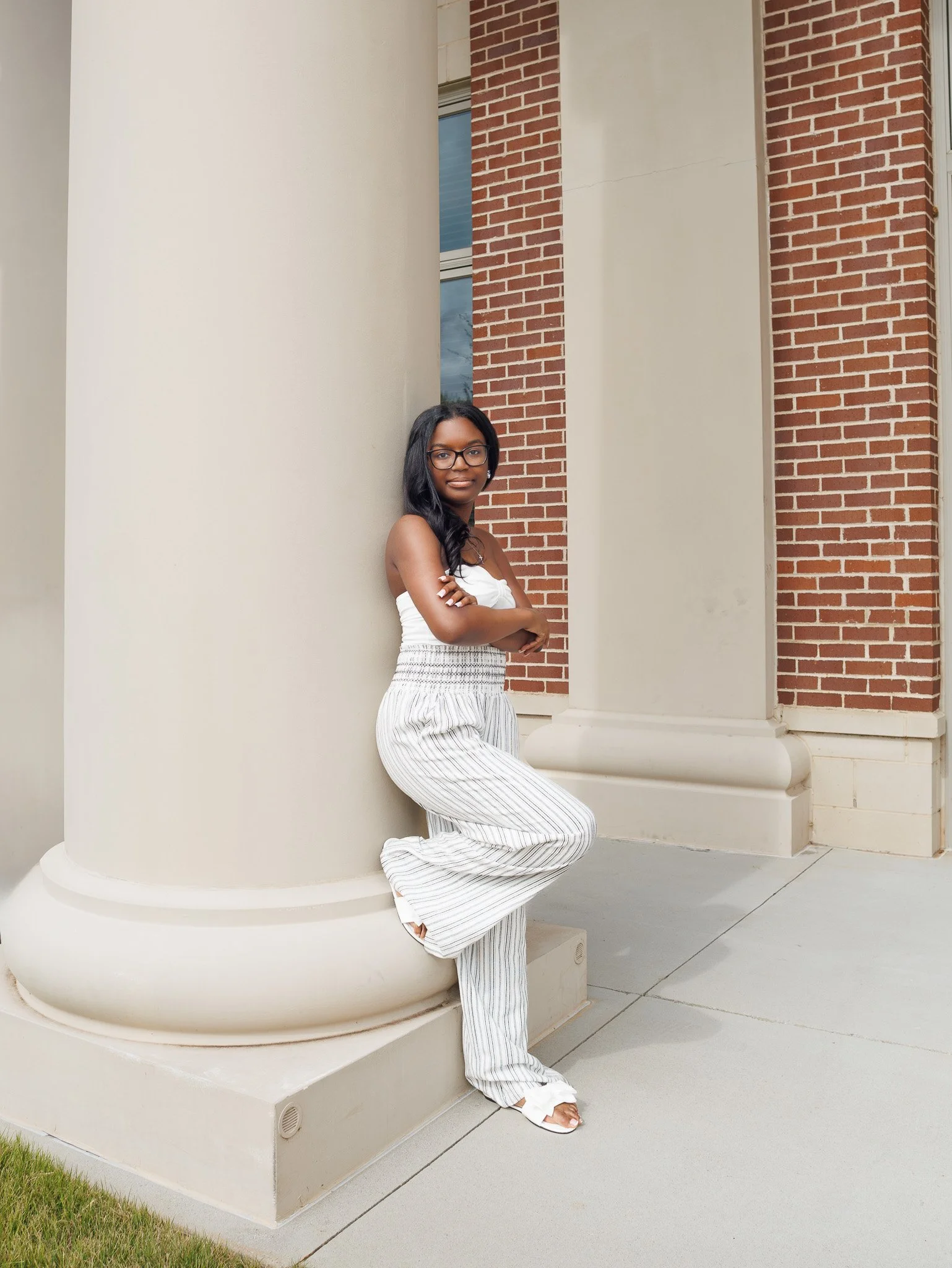 A woman leaning against a large cream-colored column outside a building with red brick walls, wearing a white strapless top and striped wide-leg pants.