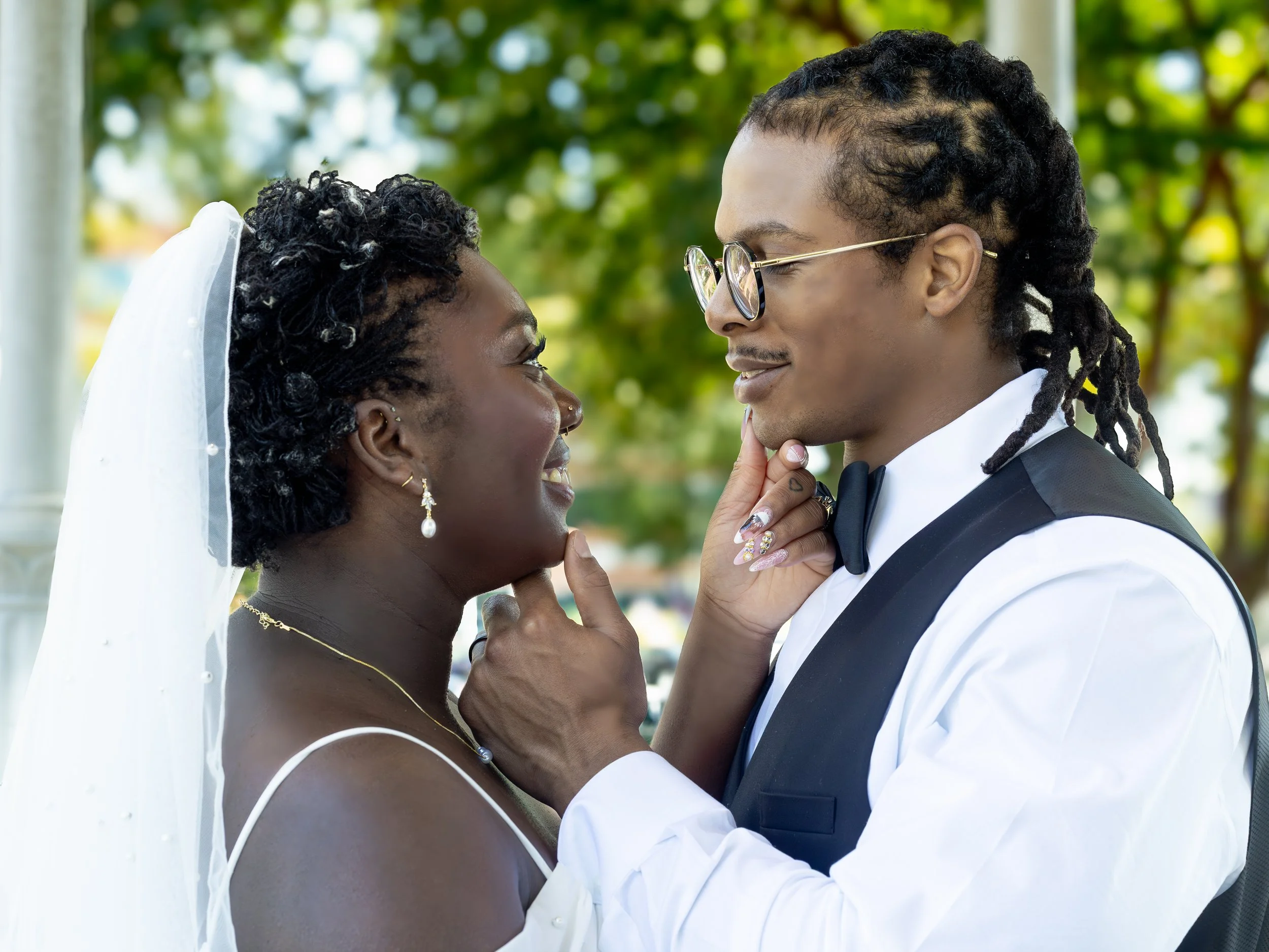 A couple dressed in wedding attire gazing into each other's eyes outdoors, with the woman in a wedding dress and veil and the man in a tuxedo with glasses.