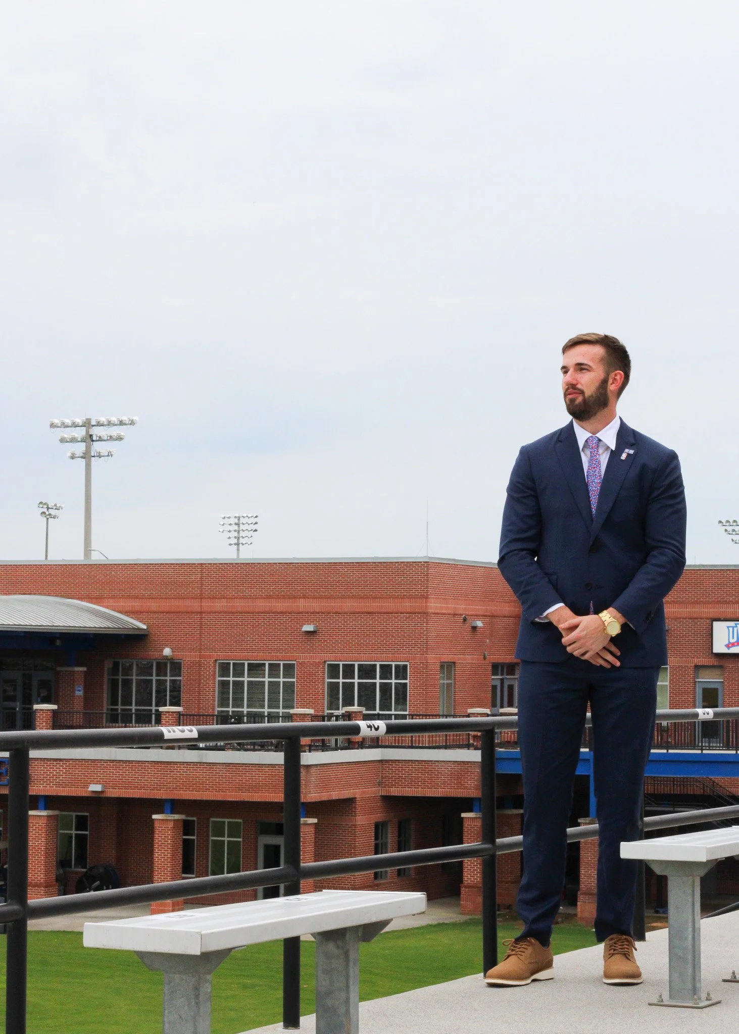 A man in a blue suit with a colorful tie and brown shoes standing on an outdoor sports stadium balcony. He has a beard, short brown hair, and is wearing a gold watch. The background shows a red brick building, field, and multiple stadium lights again