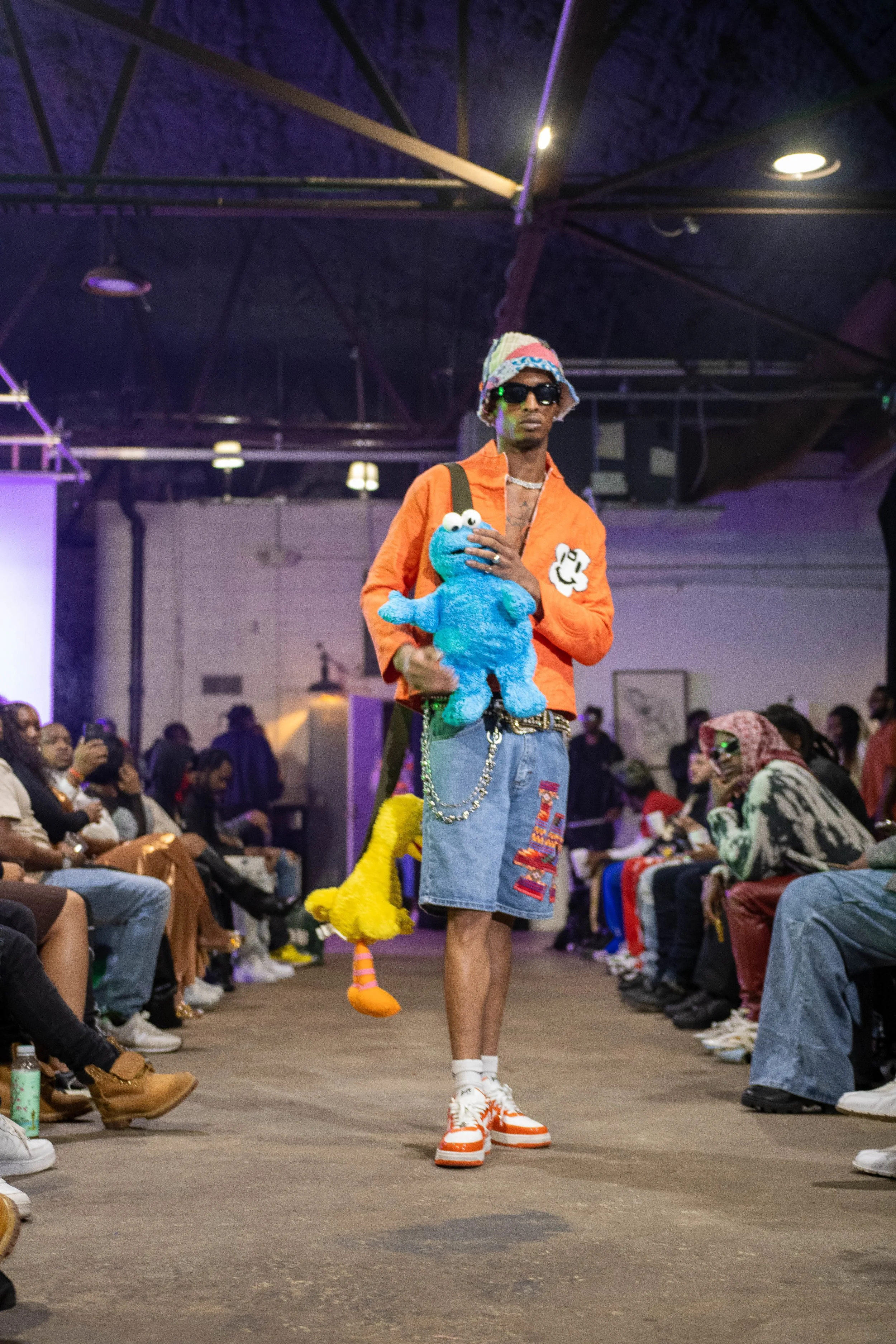 Man walking on runway holding a Barney plush toy, wearing an orange jacket, denim shorts, sneakers, sunglasses, and a multicolored knit hat, with an audience watching.