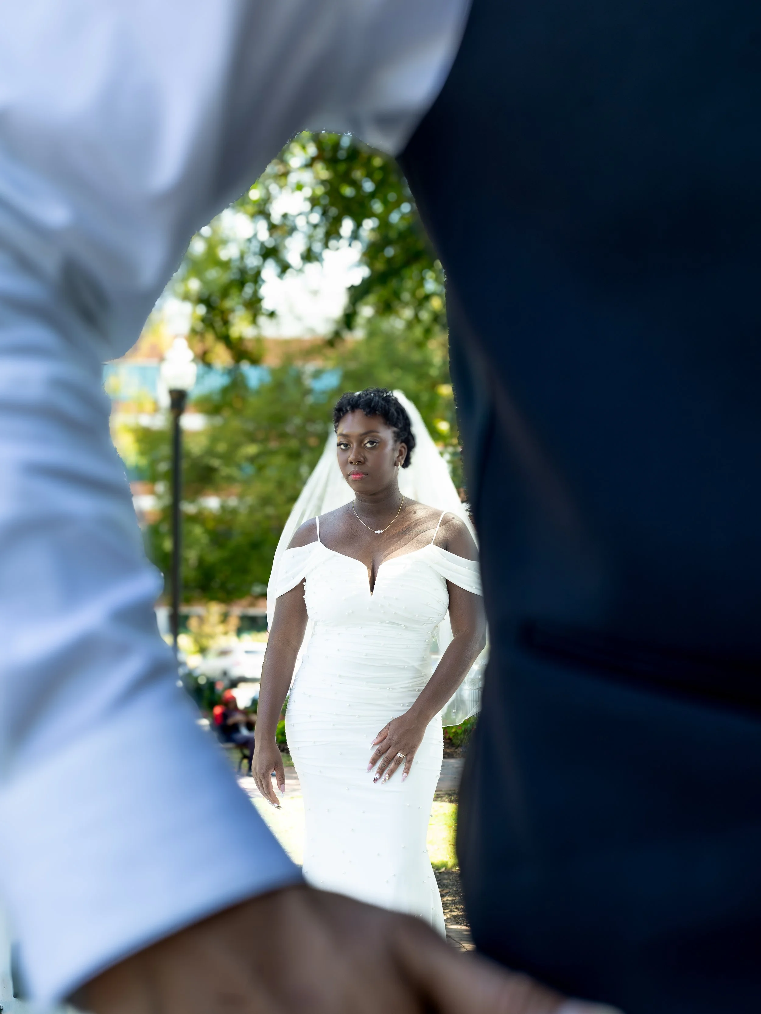 A bride in a white wedding dress standing outdoors, framed by the backs of two people in wedding attire.