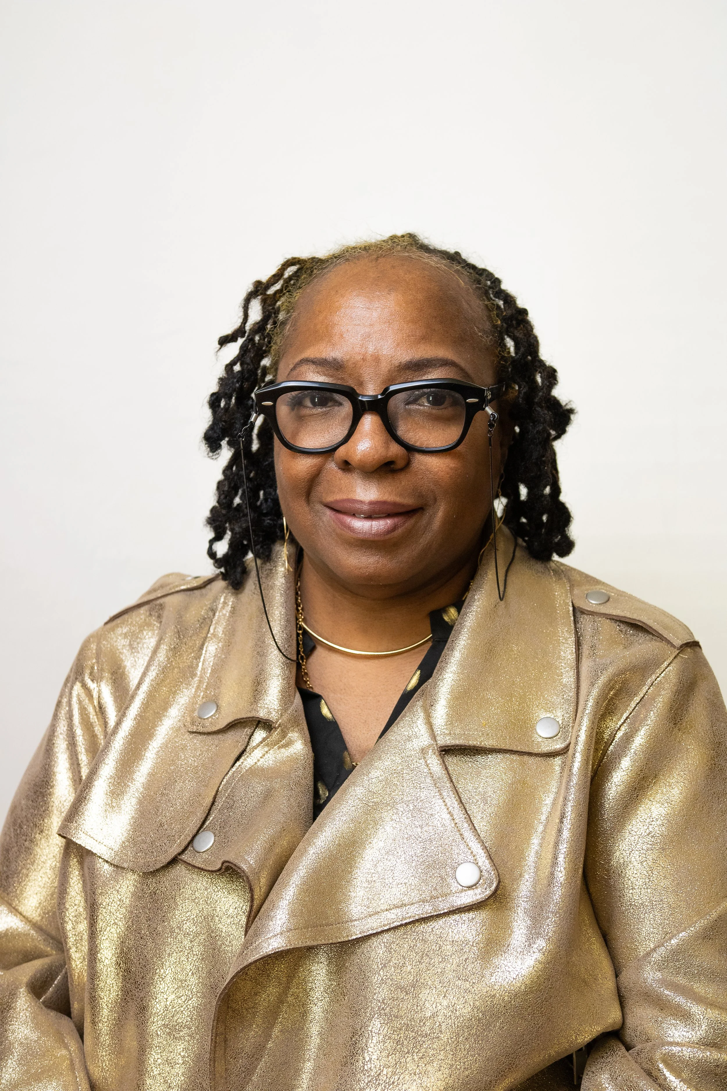A woman with glasses, gold earrings, and a gold jacket smiling at the camera.