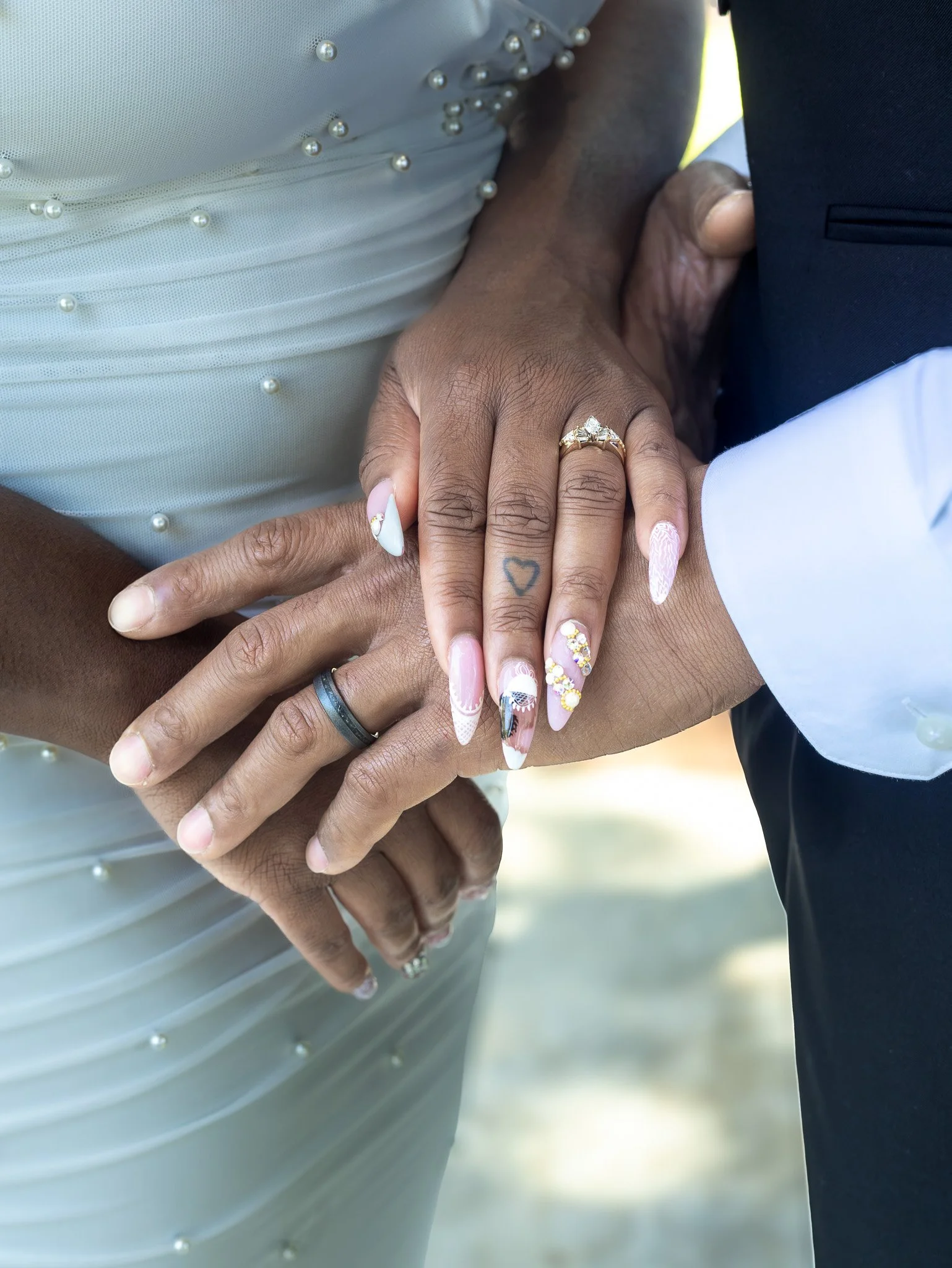 Close-up of two people holding hands, showing wedding rings and detailed nails, with part of wedding dress and suit visible.