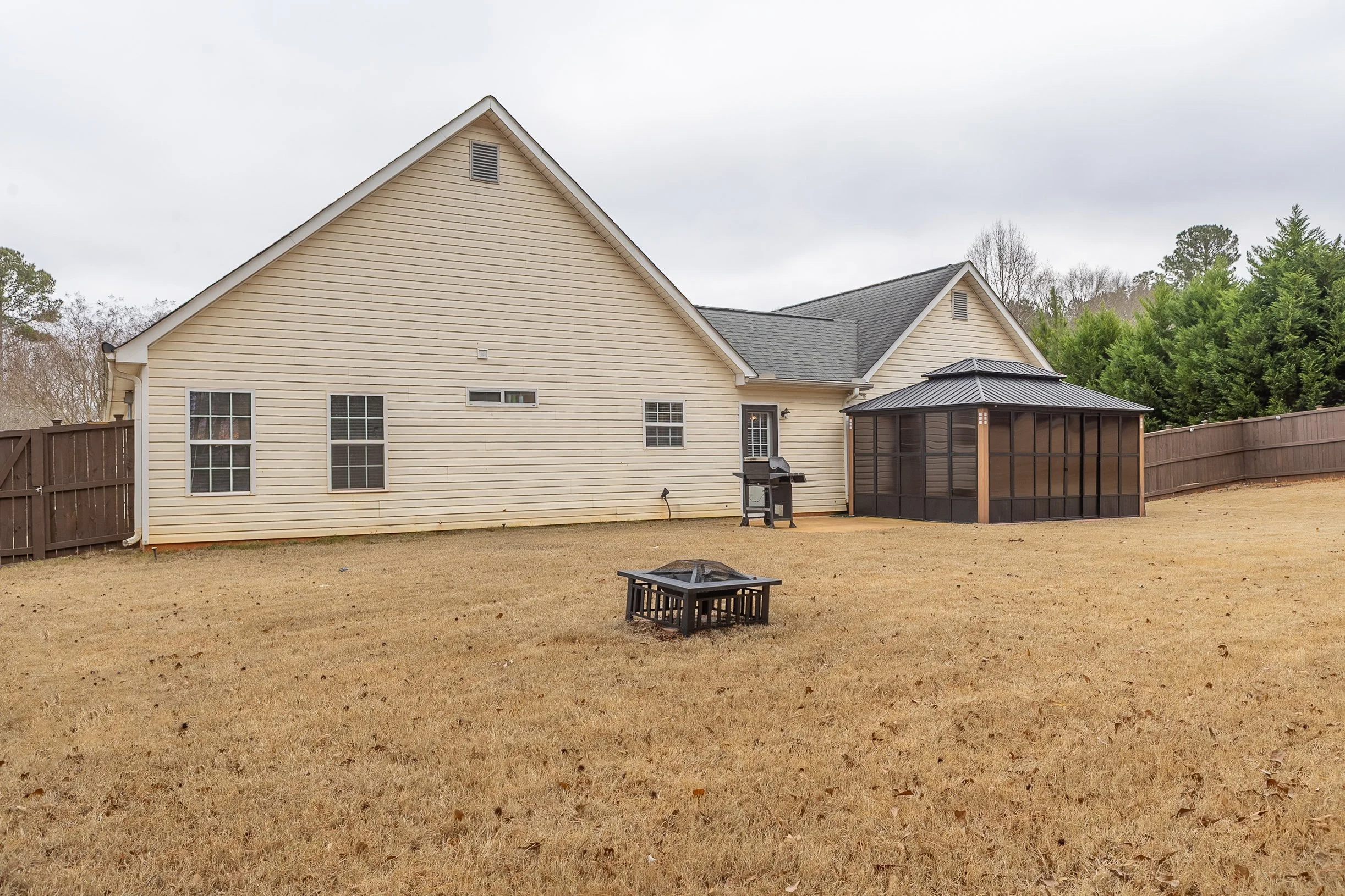 Backyard of a house with beige siding, fenced yard, grill, fire pit, and screened porch, with trees in the background.