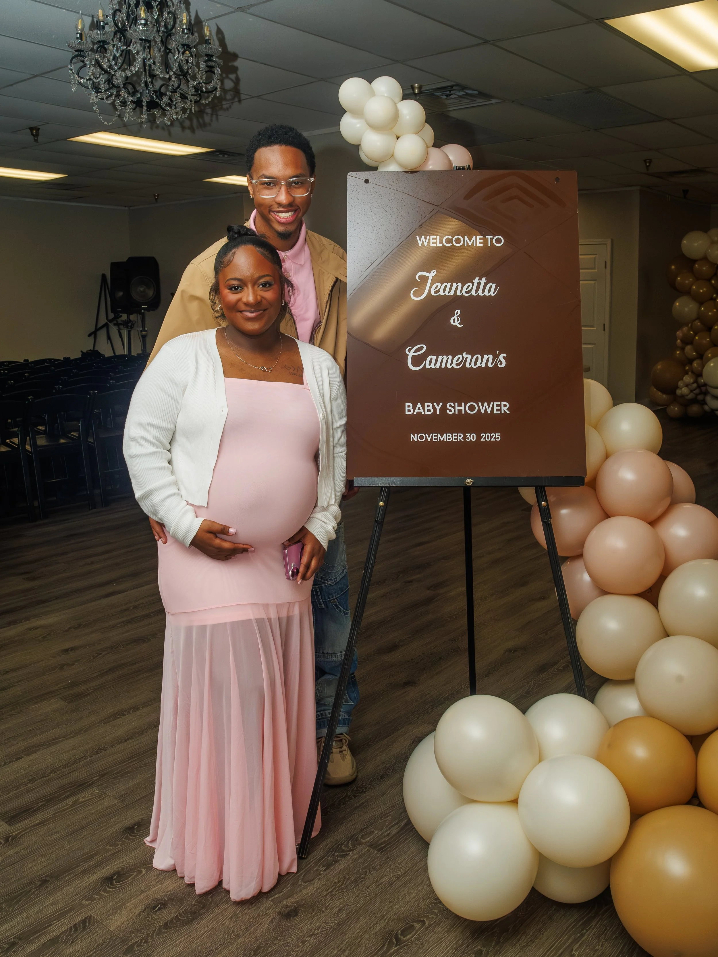 A pregnant woman dressed in a pink dress and white cardigan standing next to a man in a beige jacket and glasses, both smiling, at a baby shower event with a sign that reads 'Welcome to Jeanetta & Cameron's Baby Shower, November 30, 2025,' surrounded