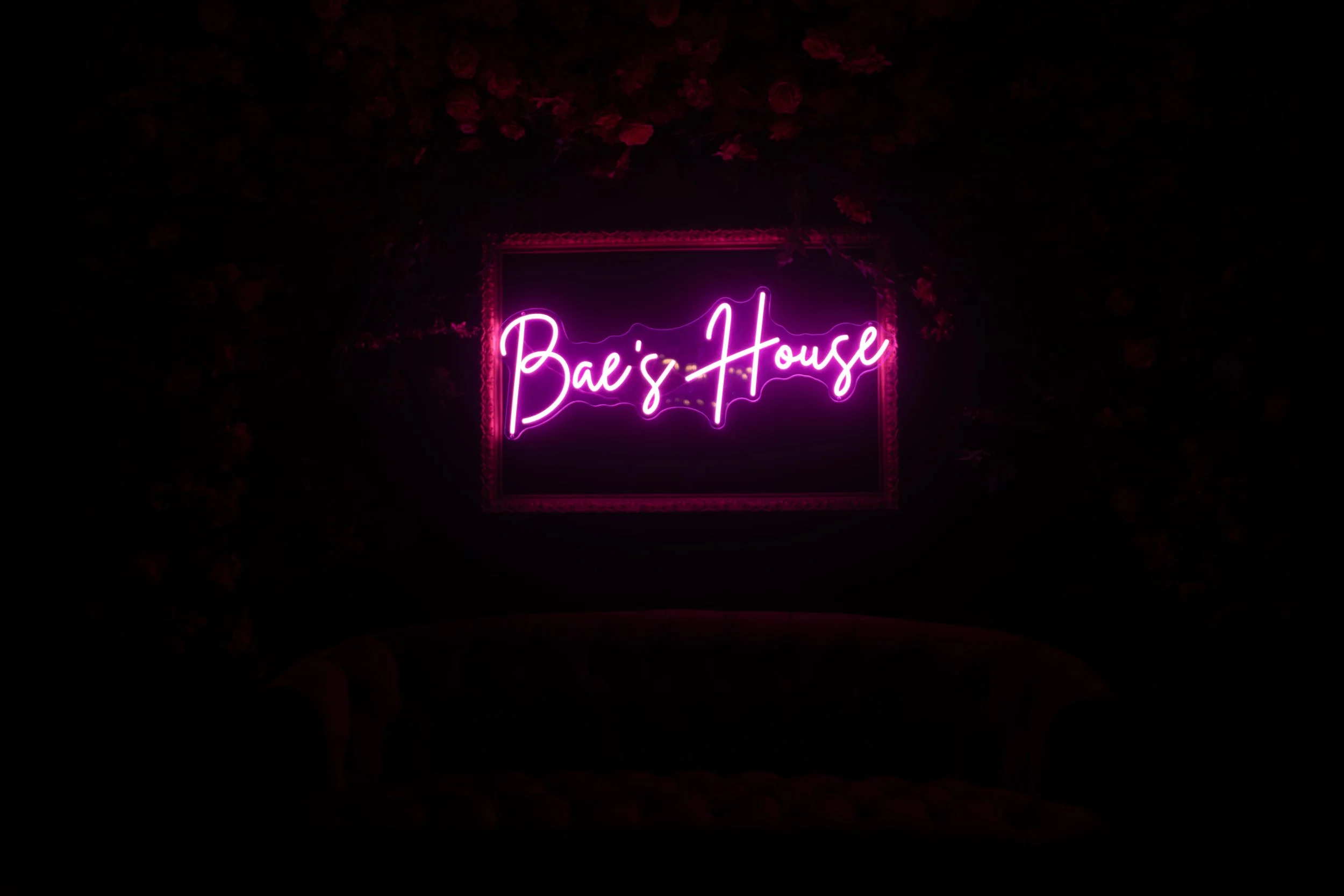 Pink neon sign that says 'Bae's House' mounted on a dark wall with floral decoration.
