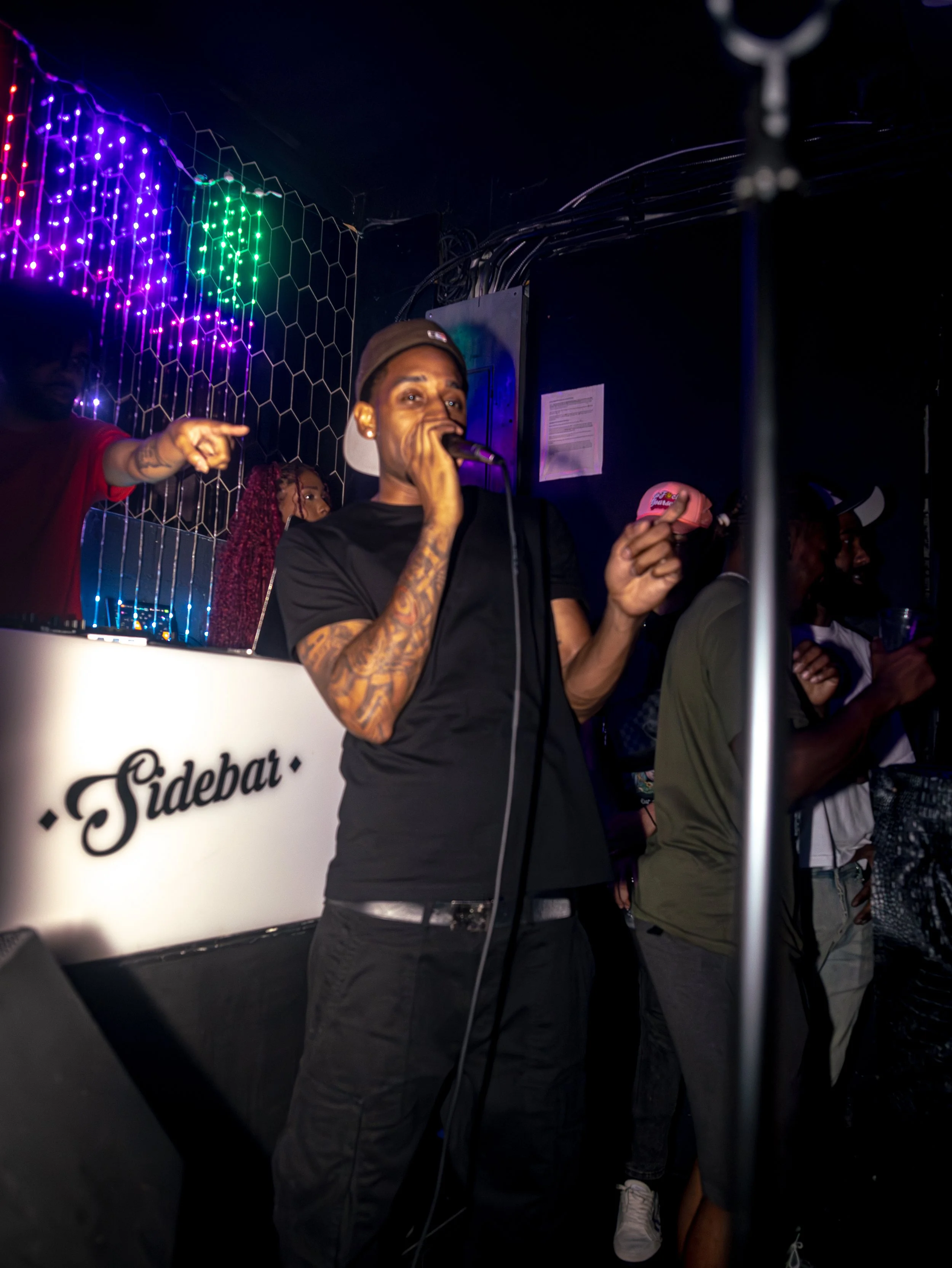 A male singer with tattoos on his arms, wearing a black t-shirt and a backwards baseball cap, holding a microphone and performing on stage at a nightclub with purple and green lights.