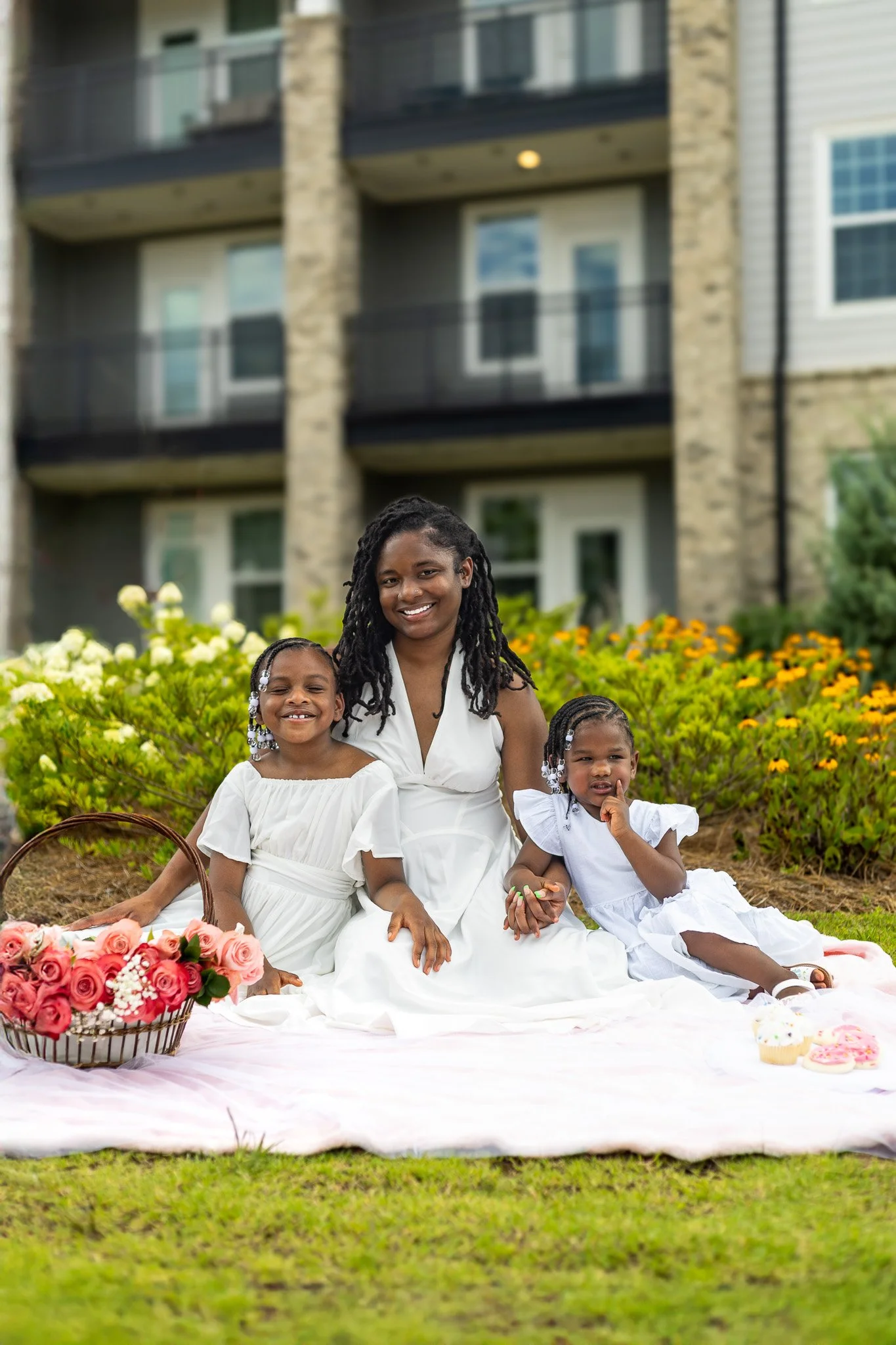 A woman and two young girls sitting on a pink blanket outdoors in front of bushes with yellow and white flowers, with a residential apartment building in the background. The woman and girls are dressed in white, and there is a basket of pink roses an