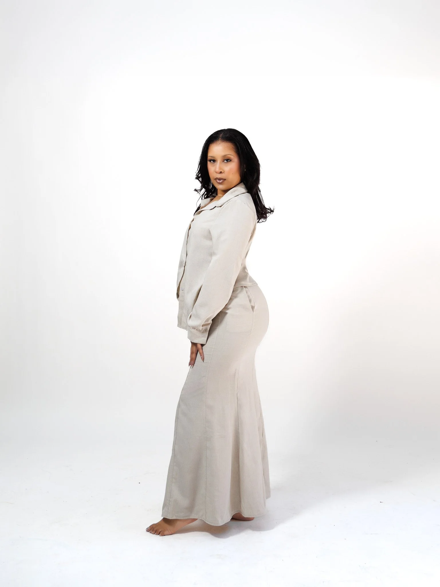 Woman with dark hair wearing a beige button-up shirt and matching wide-leg pants standing against a white background.
