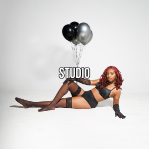 A woman with curly red hair in black lingerie and fishnet gloves posing on the floor with black and silver balloons against a white background.