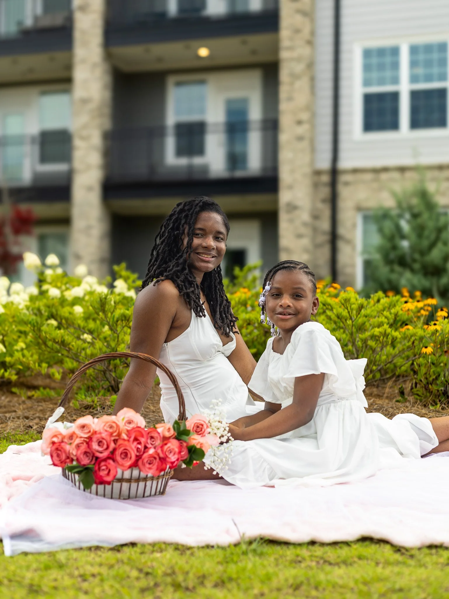 A woman and young girl sitting on a pink blanket outdoors with a basket of pink roses, in front of a building with stone and siding exterior and green bushes.