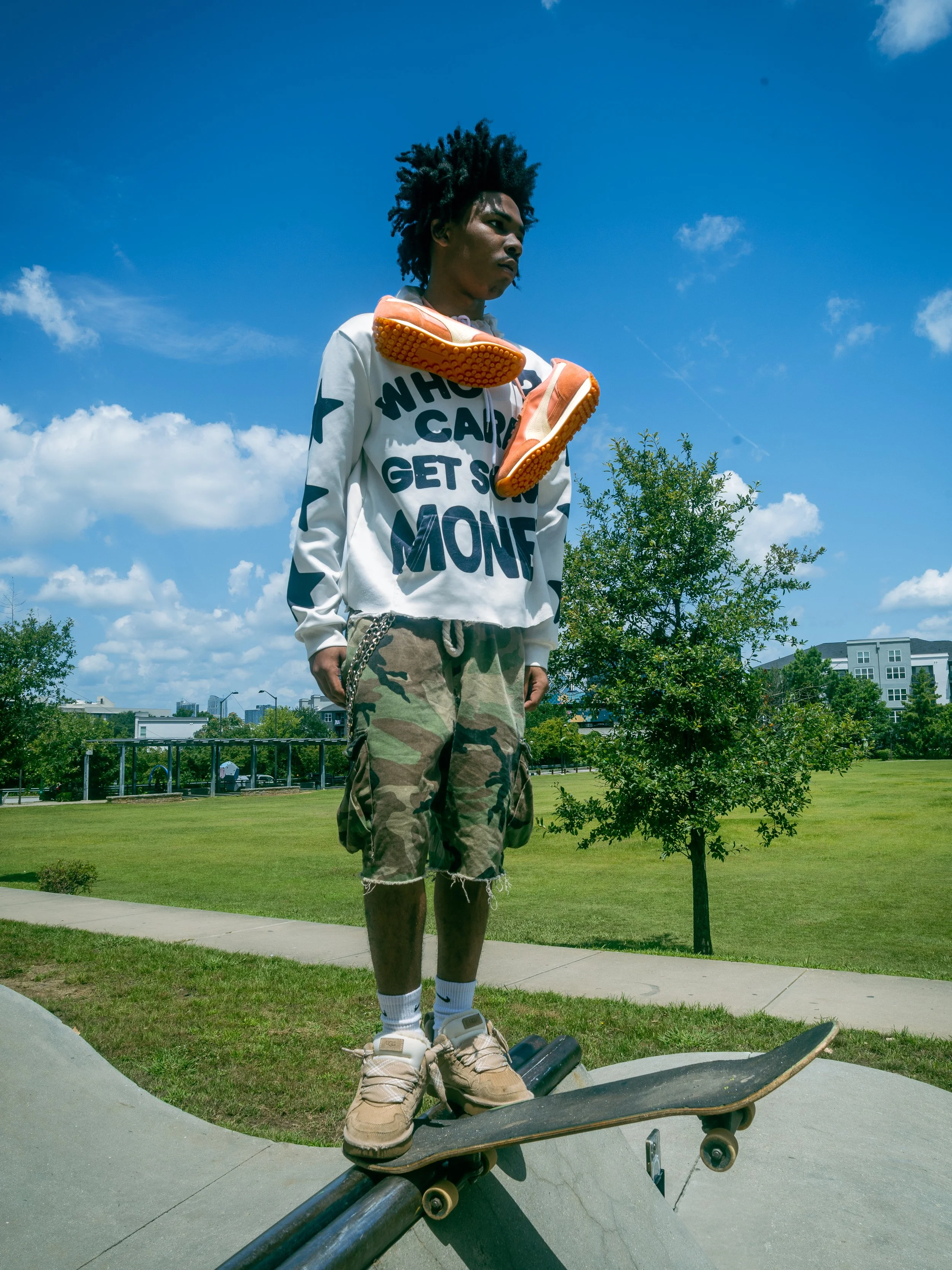 A young man with curly hair standing on a skateboard at a park on a sunny day, wearing a white sweatshirt with text, camouflage shorts, and beige sneakers, with orange shoes hanging on his shoulders.