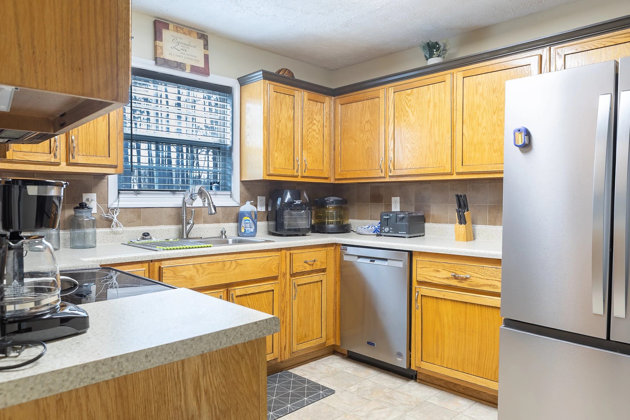 Kitchen with wooden cabinets, stainless steel refrigerator, dishwasher, countertop appliances, window, and various kitchen items.