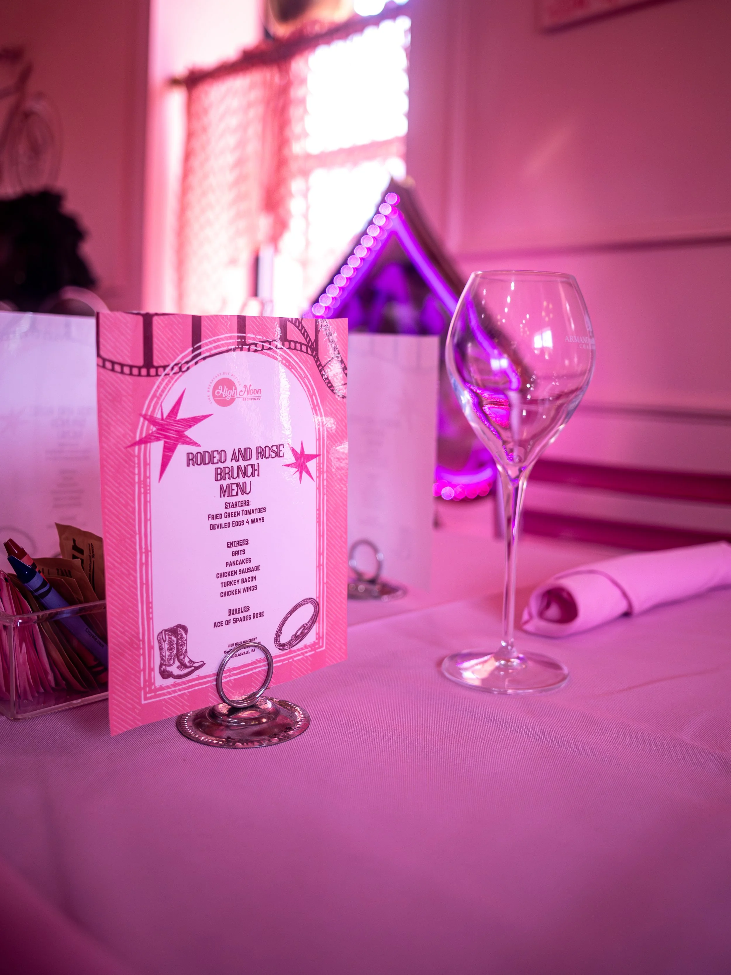 A table setting with a pink 'Rodeo and Rose' brunch menu, an empty wine glass, and a pink napkin in a pink-lit room with decorative lighting and pink curtains.