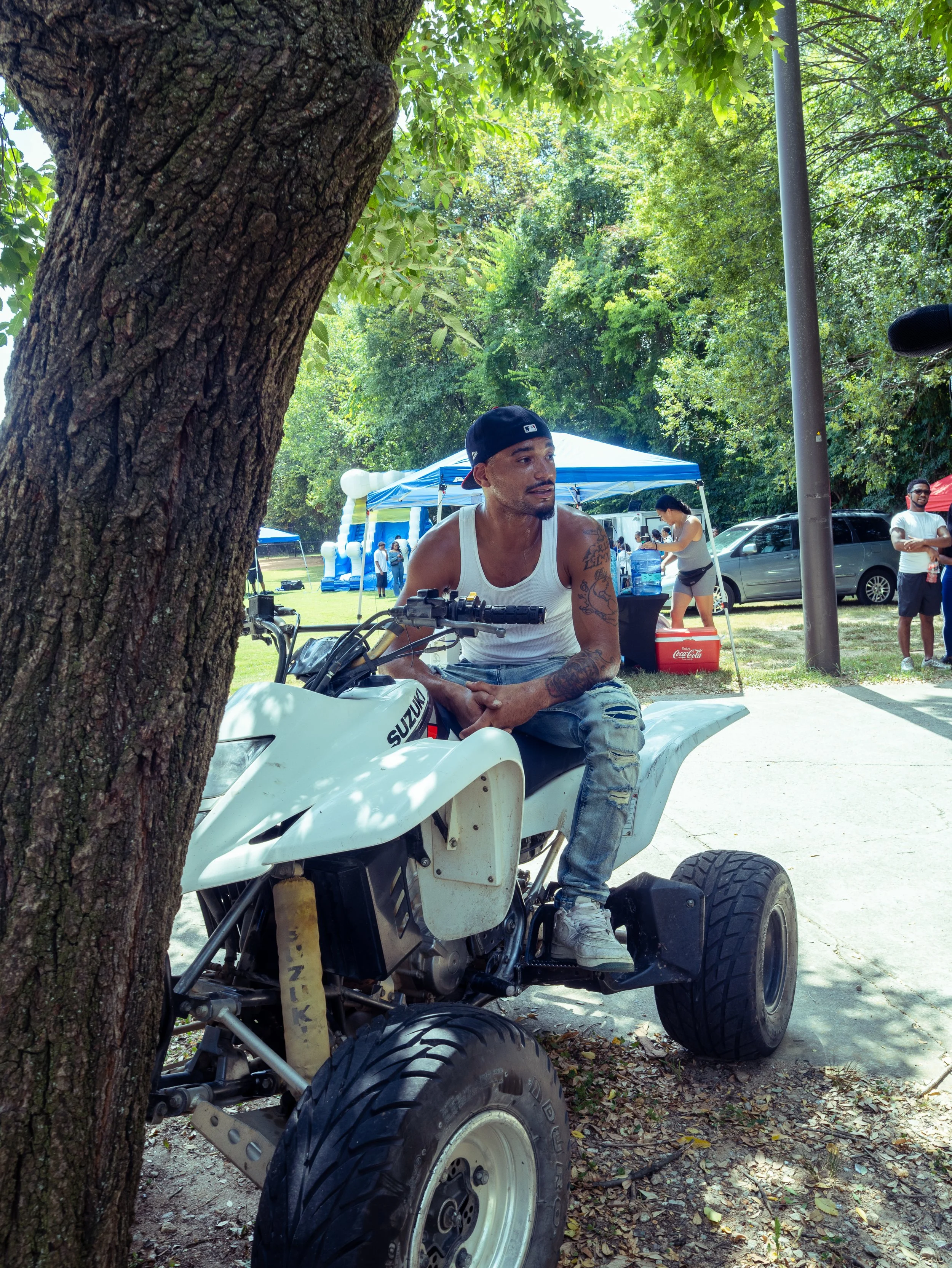 A man sitting on a white Suzuki ATV in an outdoor park setting with trees, tents, and people in the background.