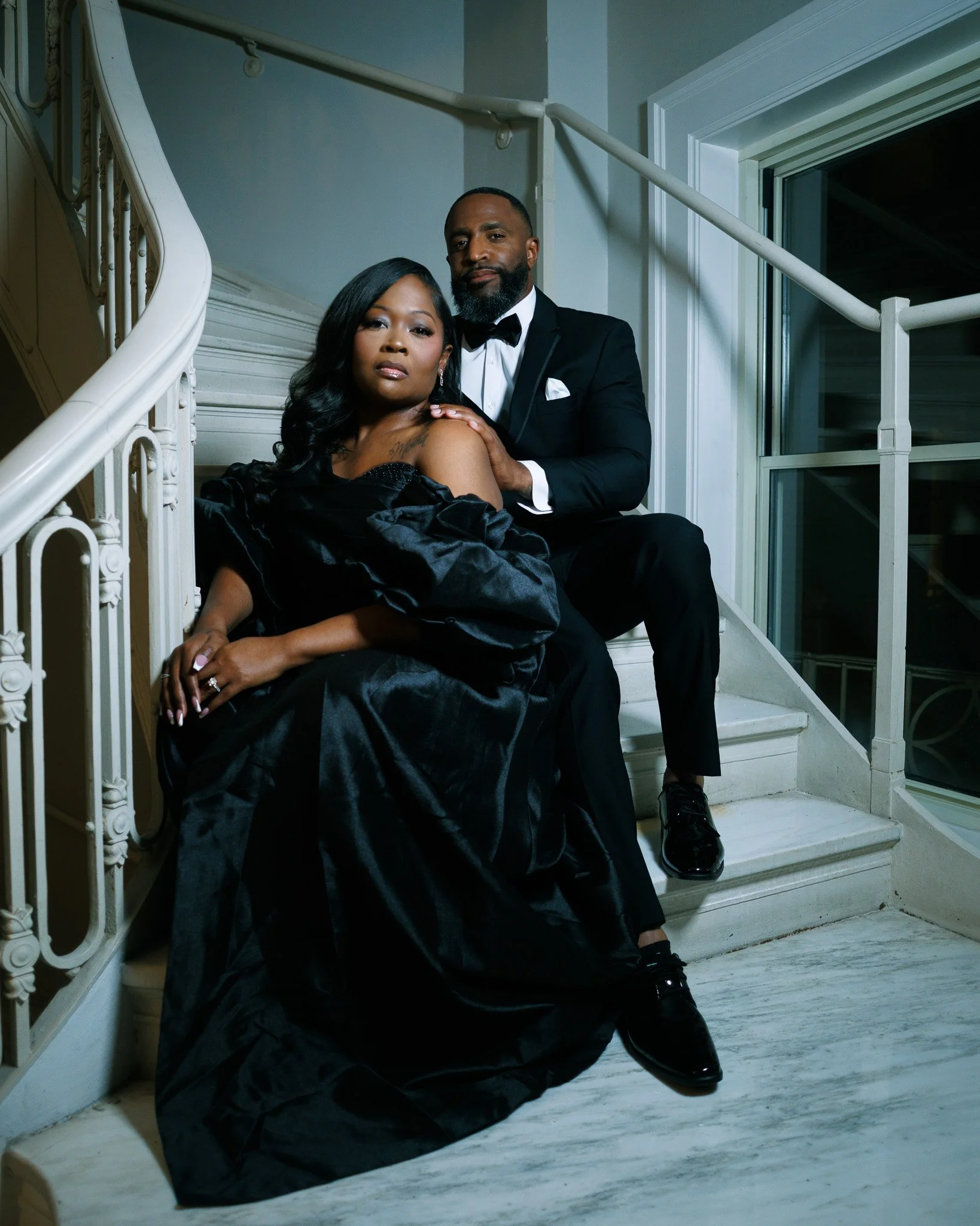 A stylishly dressed man and woman sitting on a staircase in a luxurious black tuxedo and gown.