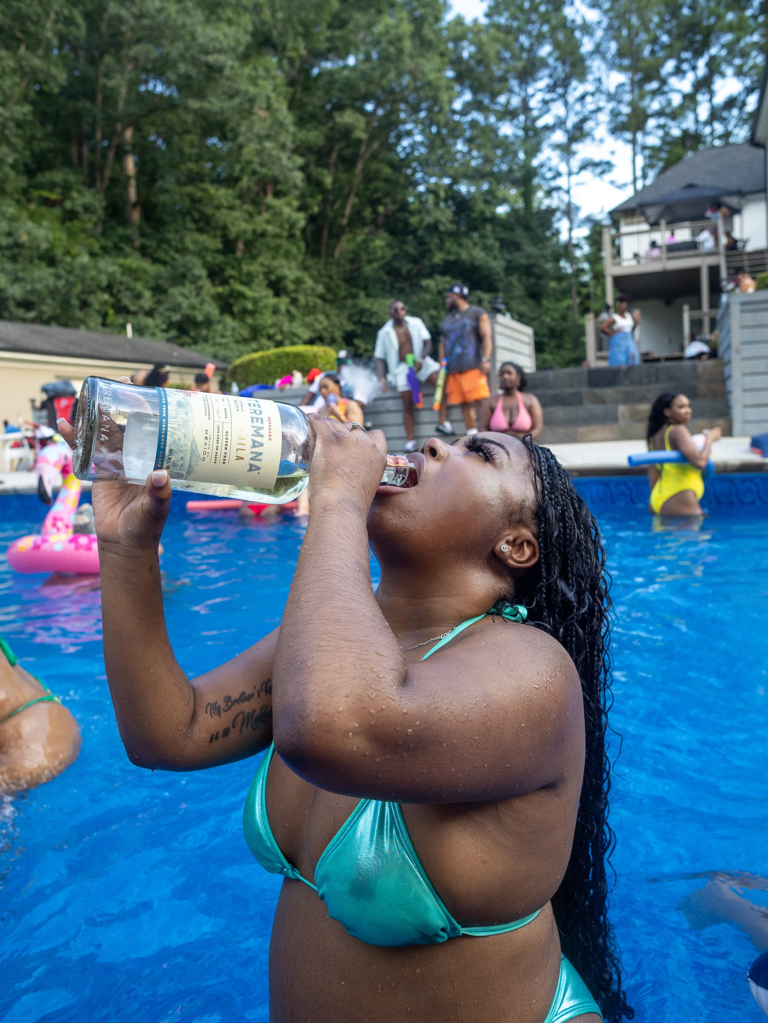 Woman in a turquoise bikini drinking from a bottle at a pool party with people on the deck and in the pool in the background.