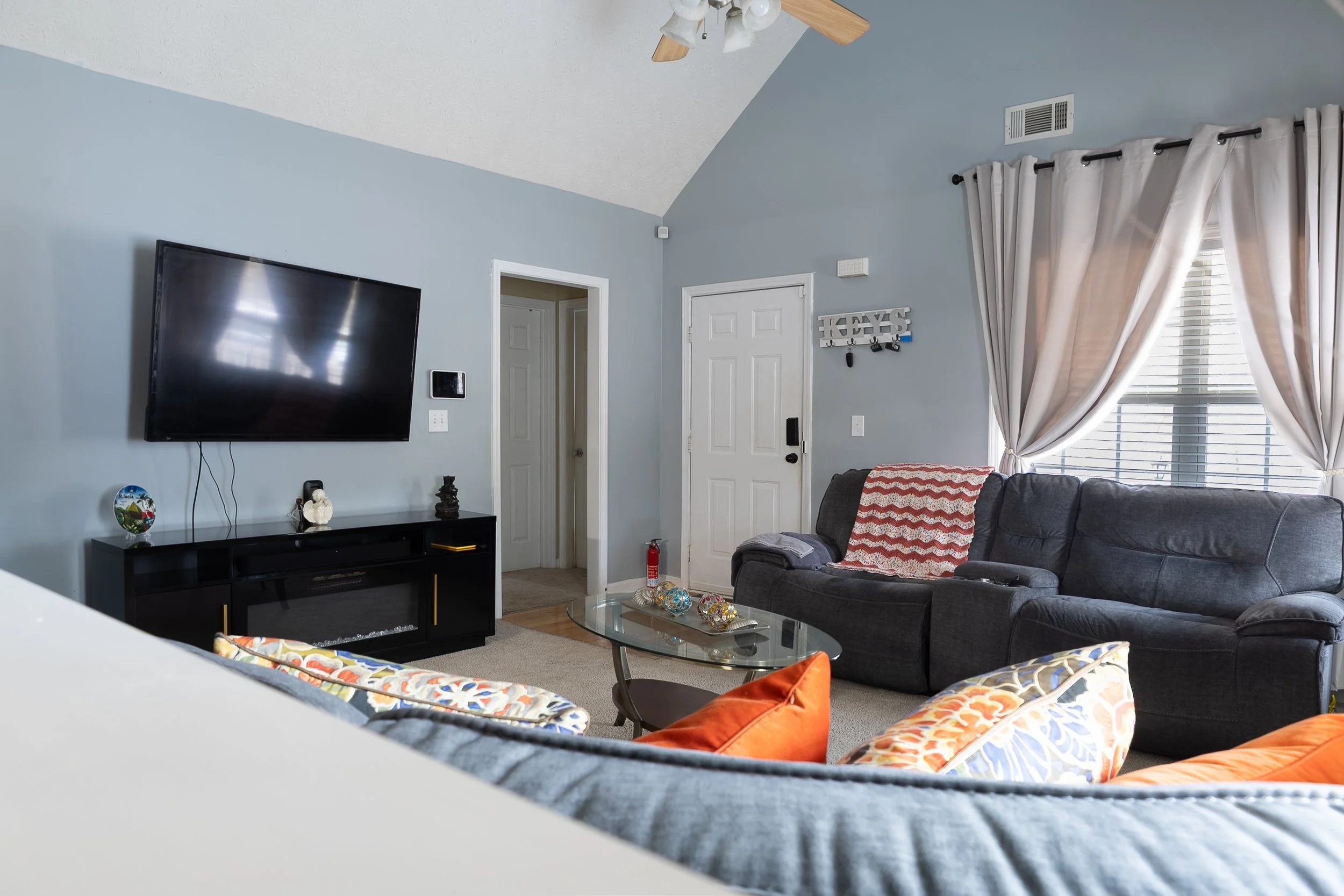 Living room with a large wall-mounted TV, a black TV stand, a gray couch with a red and white blanket, a glass coffee table with decorative items, and curtains covering a window.