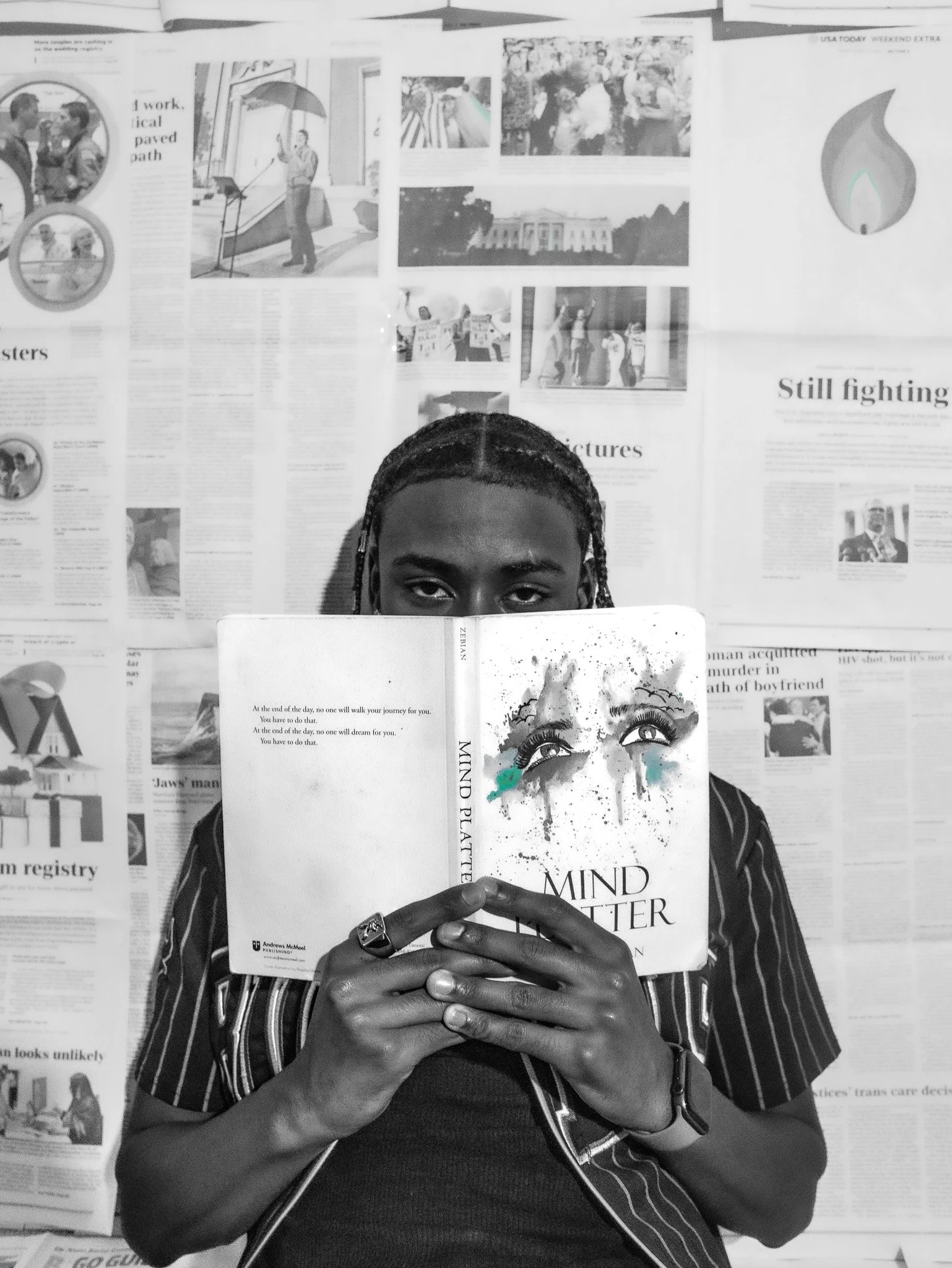 A person with braided hair is holding a book titled 'Mind Matter' in front of their face, with only their eyes visible above the book. The background is a wall covered with newspapers and photos.