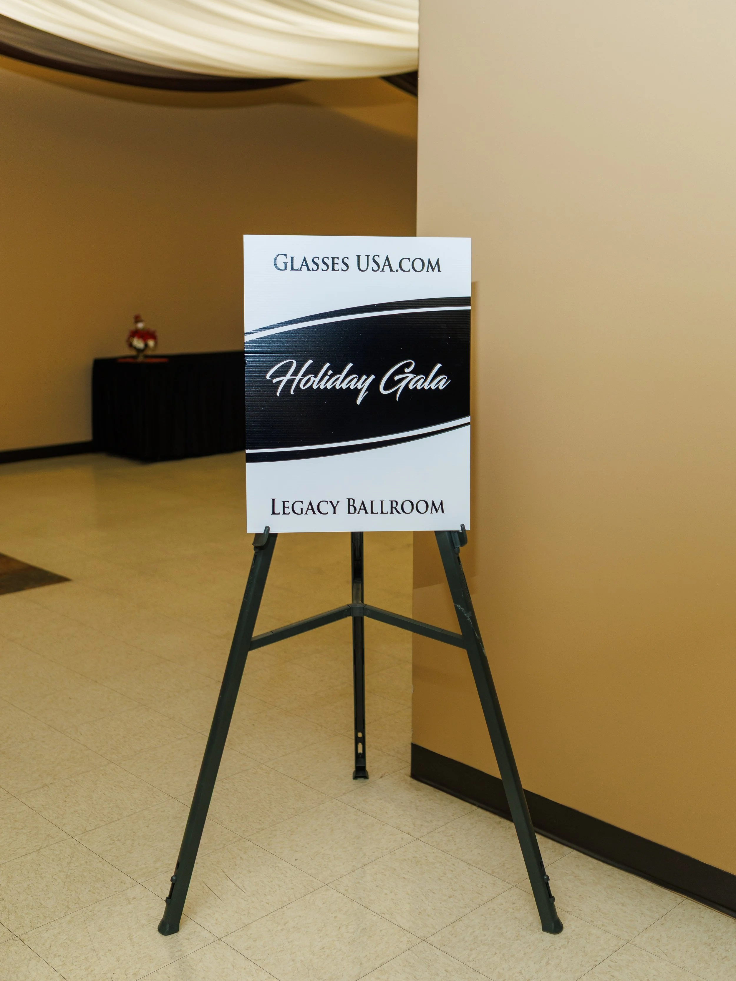 Event sign for 'Holiday Gala' at the Legacy Ballroom, organized by Glasses USA.com, on a black and white stand in an indoor venue.