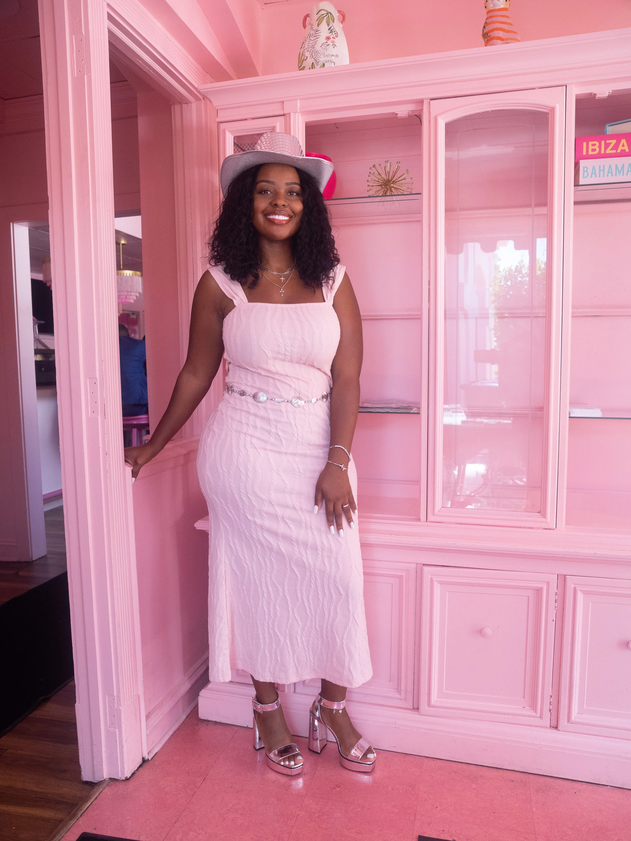 A woman dressed in a pink, sleeveless, cable-knit dress with a pearl belt, wearing silver high-heeled shoes, a hat, and jewelry, standing in front of a pink wall with decorative items and a sign that reads 'IBIZA BAHAMAS'.