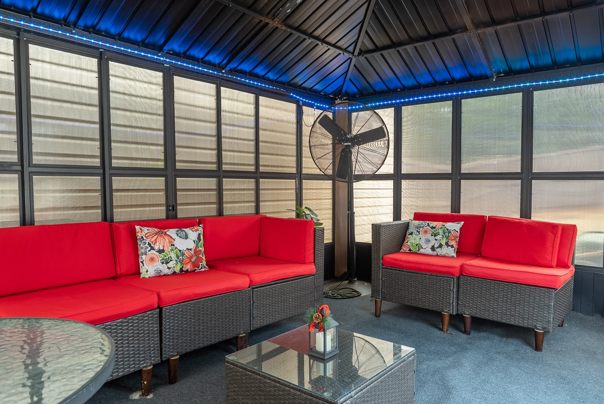 Patio enclosed porch with wicker sectional sofas with red cushions, tropical patterned throw pillows, a glass-topped side table with Christmas decor, a floor fan, and string lights on the ceiling.
