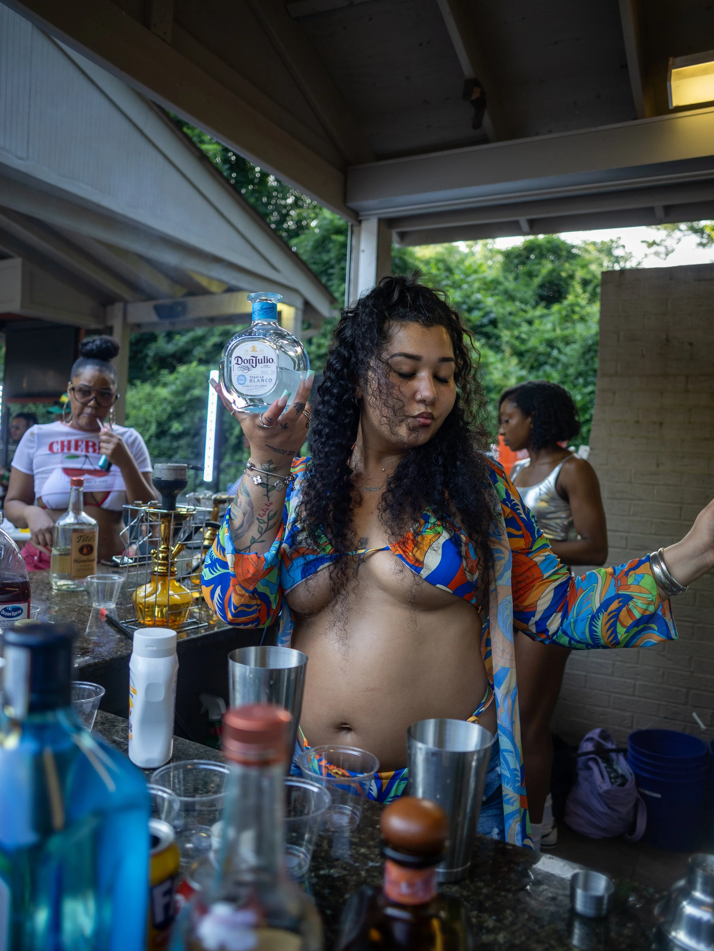 A woman with long curly hair and a colorful shirt is holding a bottle of Don Julio Tequila at an outdoor bar, with other women in the background during a gathering.
