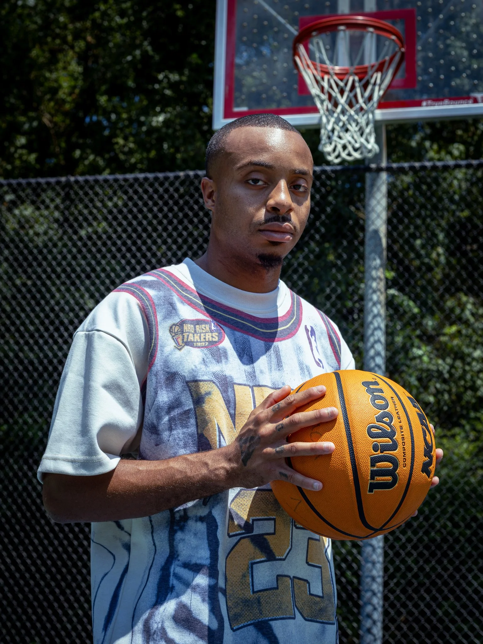 Young man with a tattooed hand holding a Wilson basketball on an outdoor basketball court.