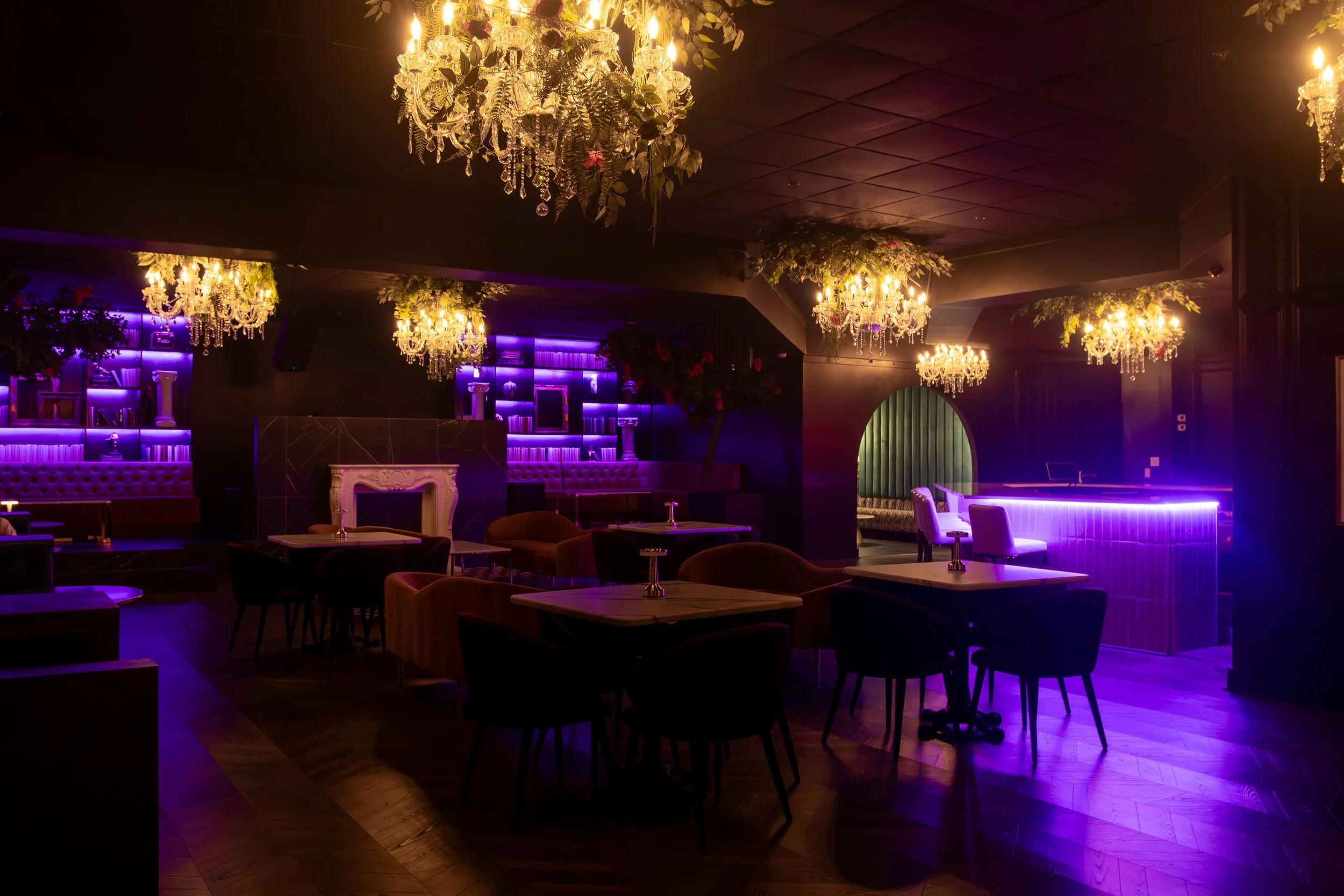 An elegant bar or lounge with purple neon lighting, chandeliers, velvet seating, and a cozy, luxurious atmosphere.