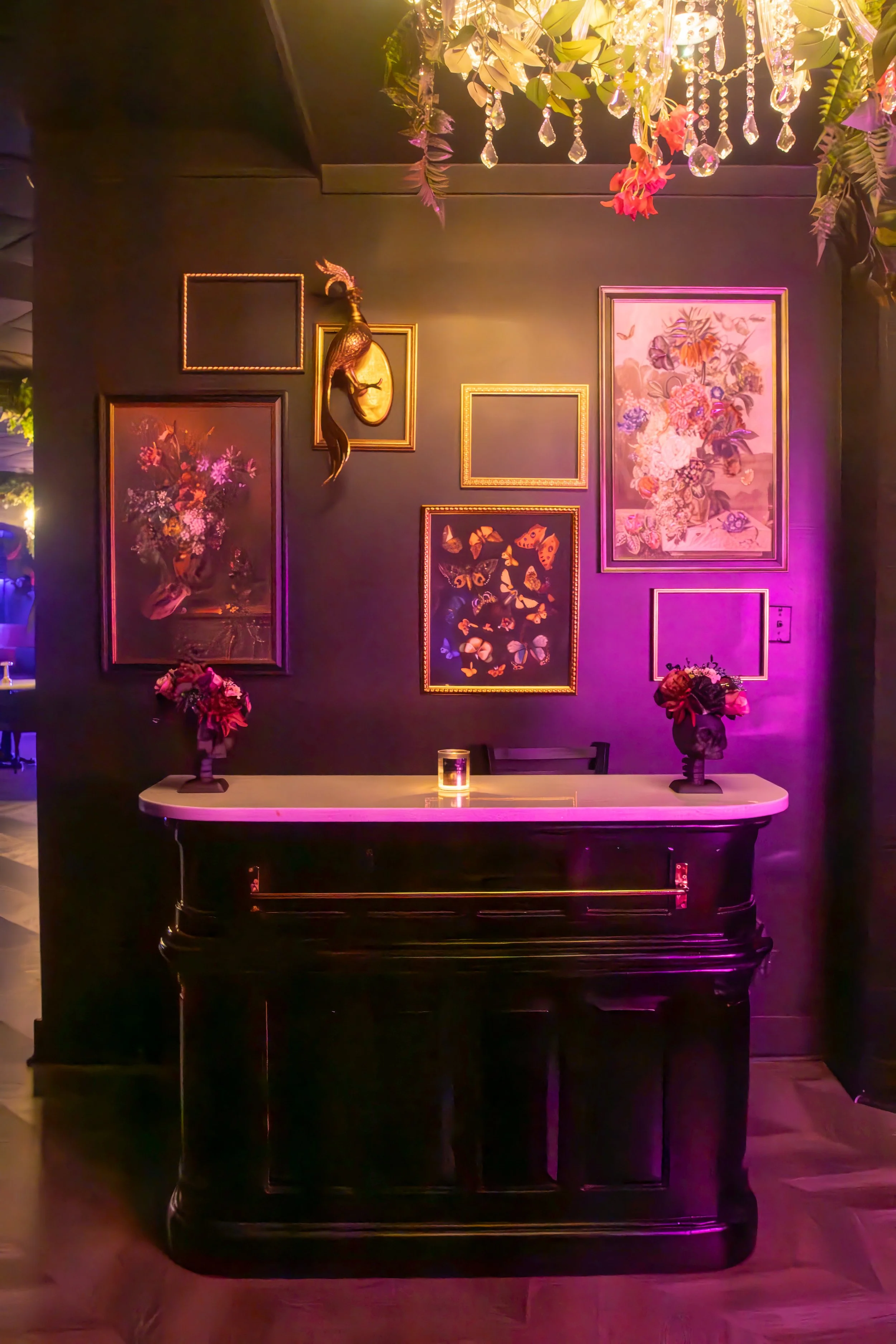 A black grand piano in front of a dark wall decorated with framed artwork, floral arrangements, and a bird sculpture, illuminated with colorful purple and pink lighting.