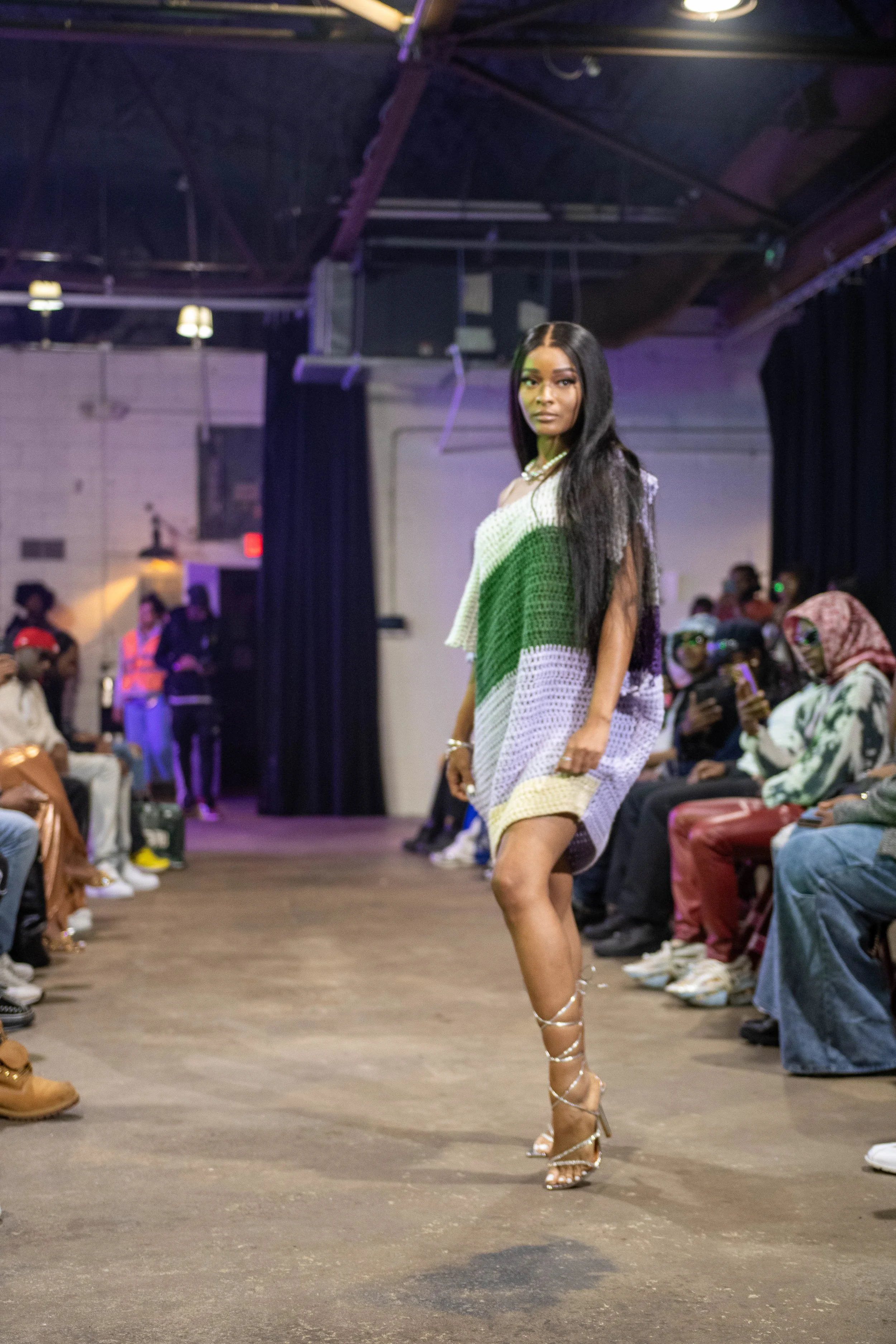 A woman walking on a runway during a fashion show, wearing a multi-colored crochet dress and high heels, with an audience seated on either side.