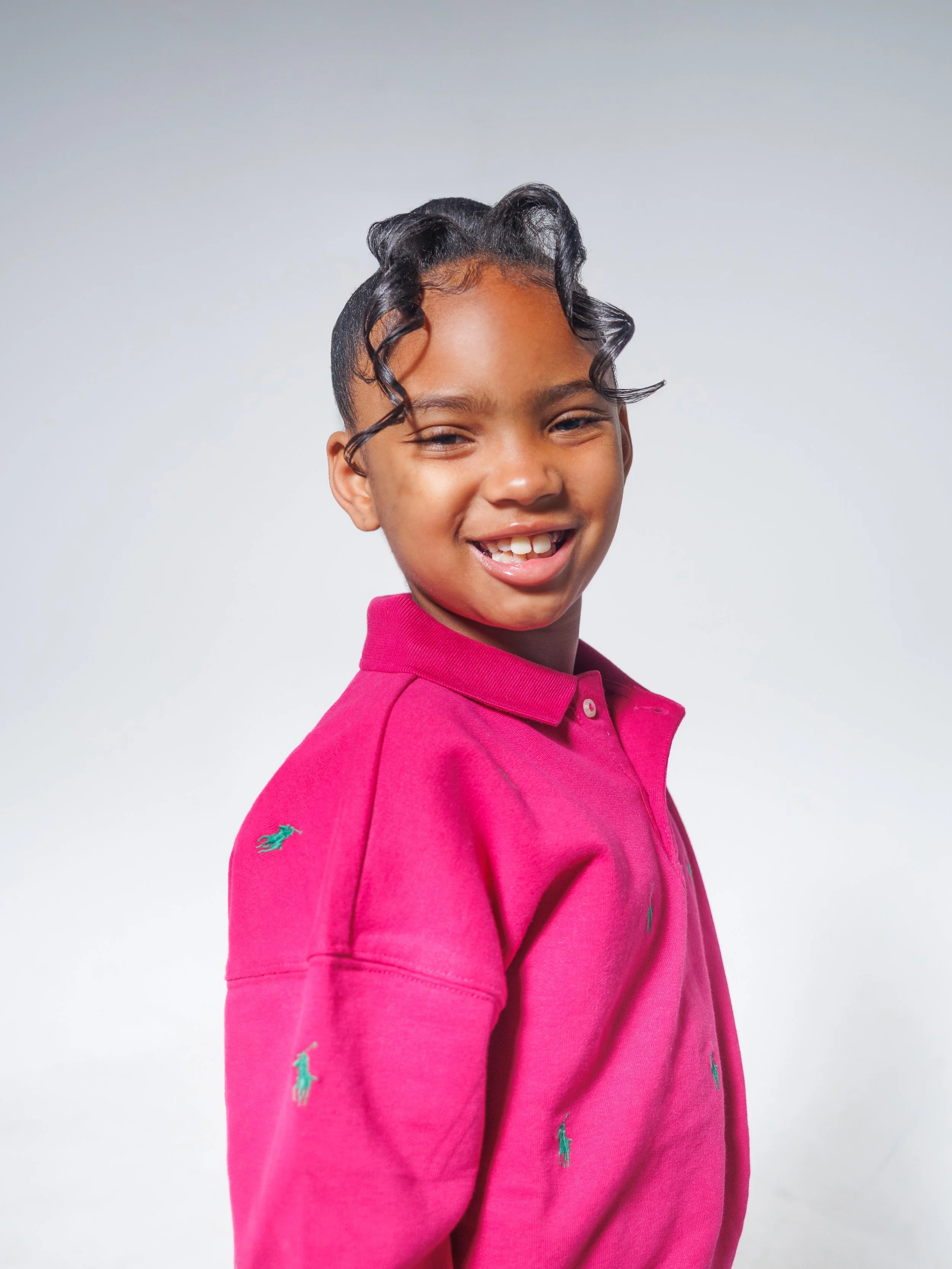 A young girl with curly hair and a smile wearing a pink polo shirt with small embroidered green logos, standing against a plain white background.